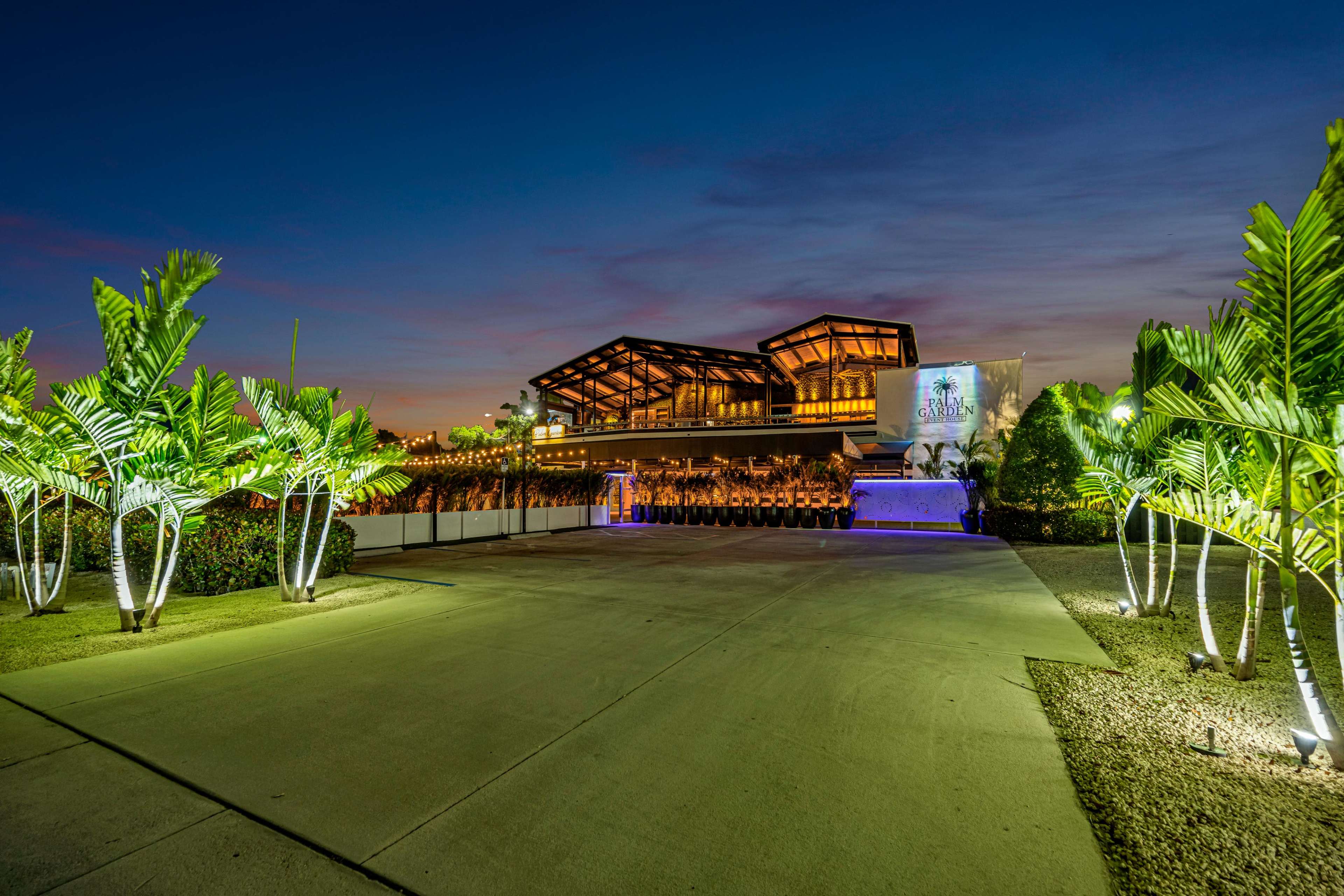 The image shows a modern, illuminated building surrounded by palm trees at dusk, with a clear driveway leading up to it.