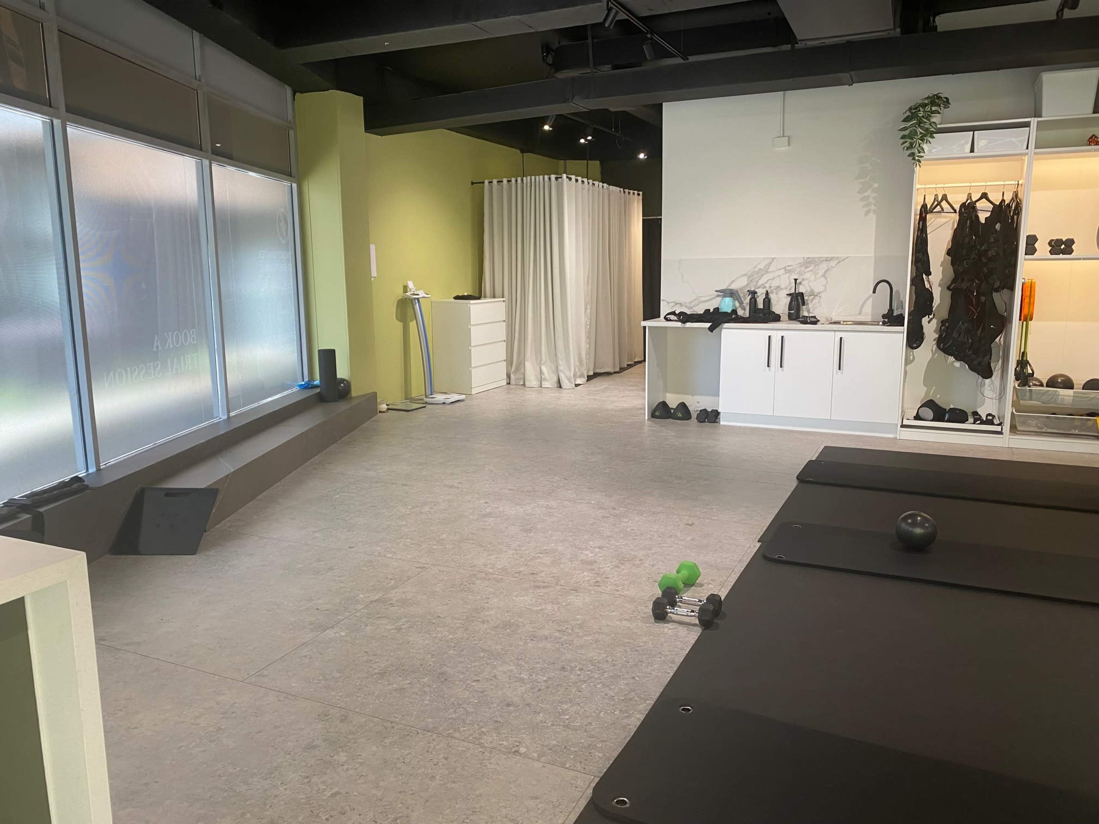 The image shows a modern fitness studio with workout mats on the floor, weights scattered around, and a bright area featuring cabinets and a curtain partition.