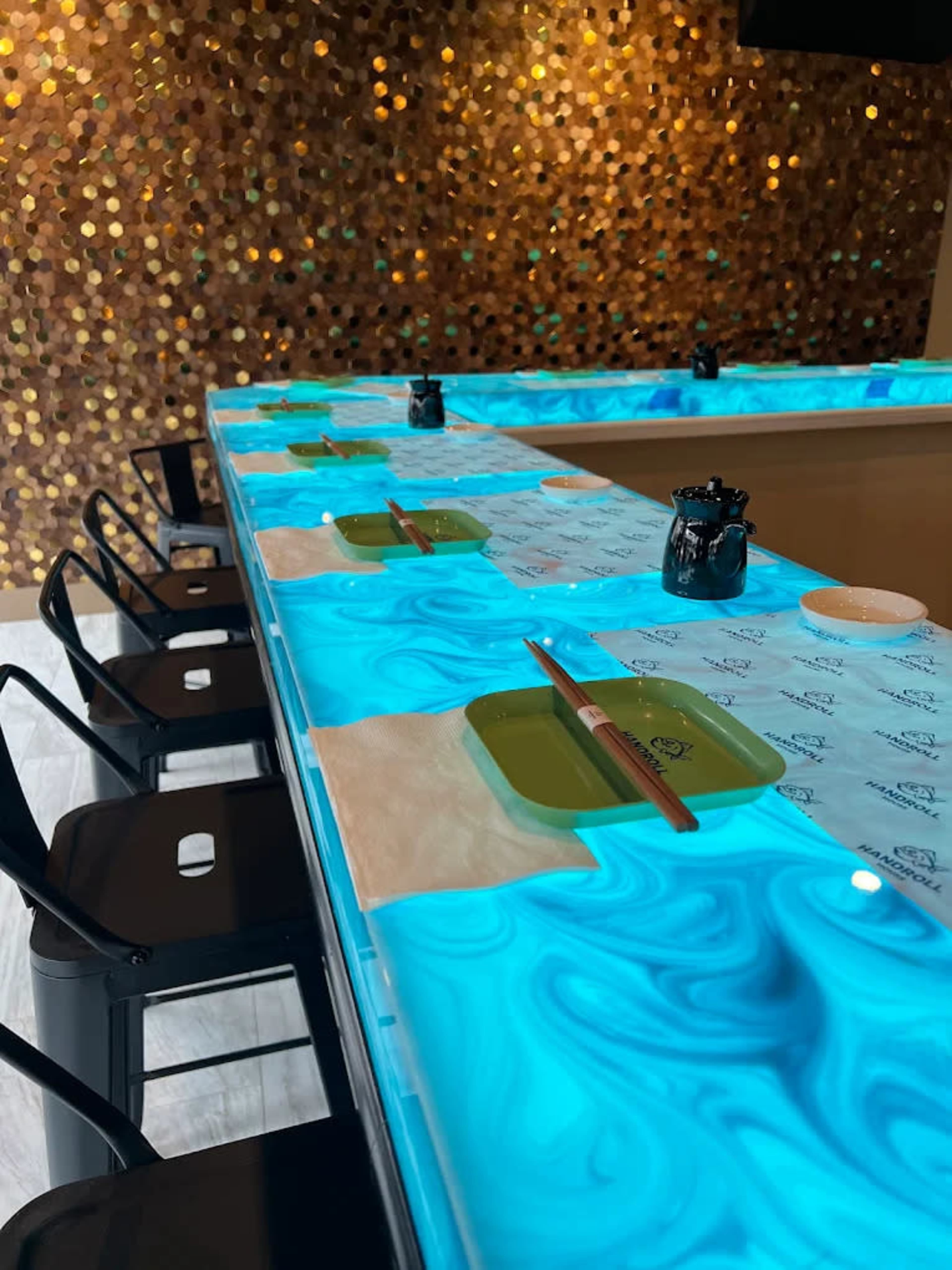 The image shows a dining counter with a luminous blue surface, neatly arranged with green plates, chopsticks, and sauce containers at each seat.