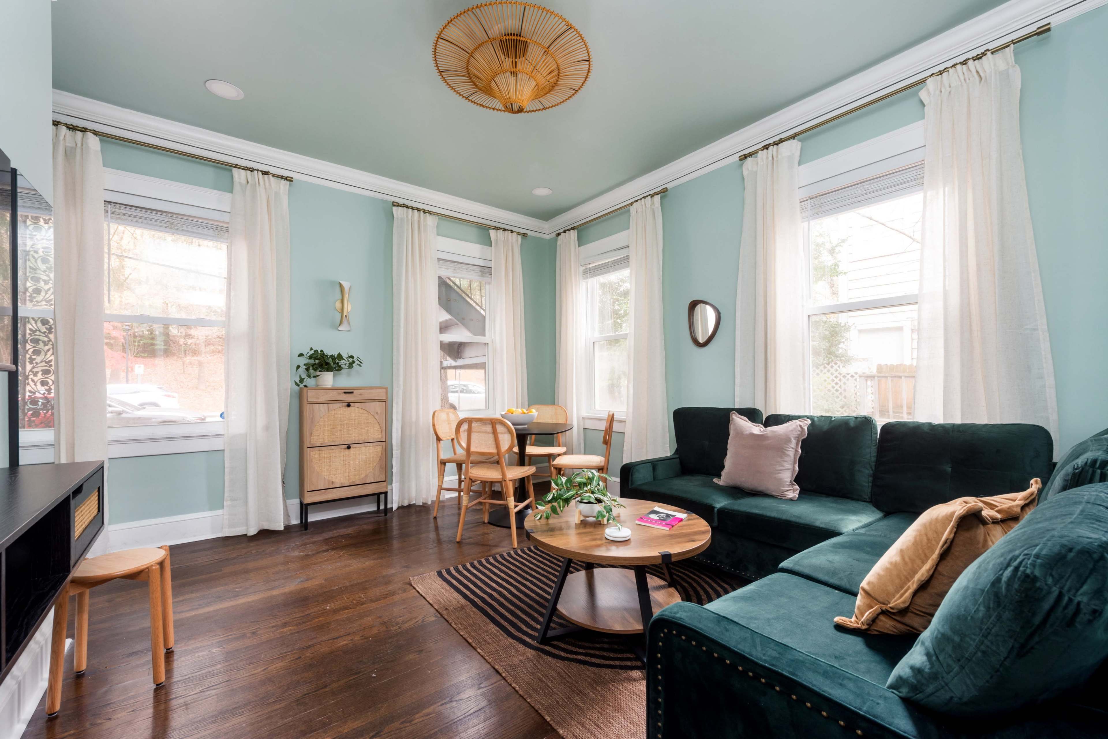 The image shows a cozy living room with a green sofa, a round coffee table, and large windows adorned with sheer curtains.