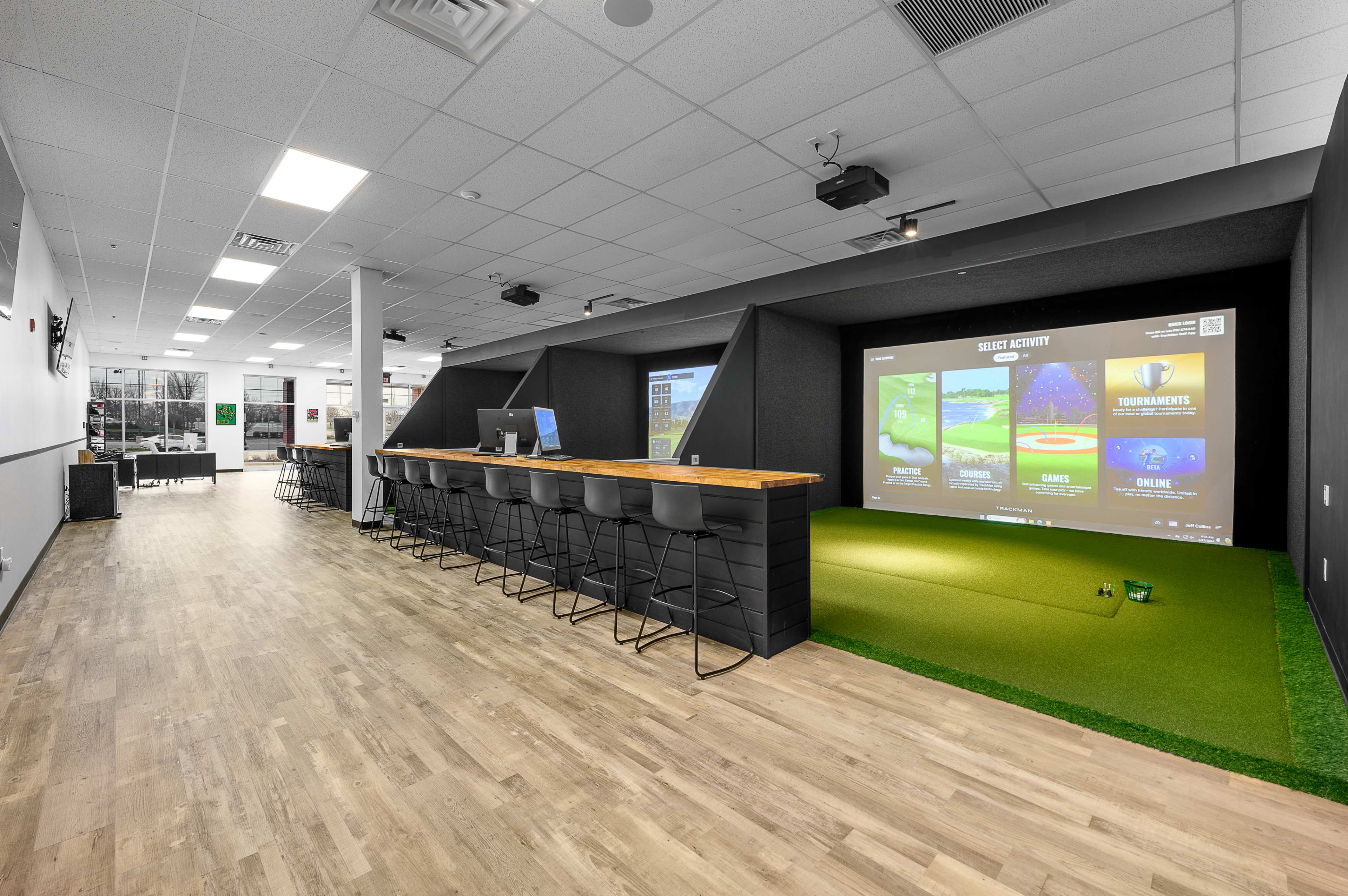 The image shows a modern indoor sports facility featuring a bar area with high stools, a large screen displaying gaming activities, and a putting green surface.