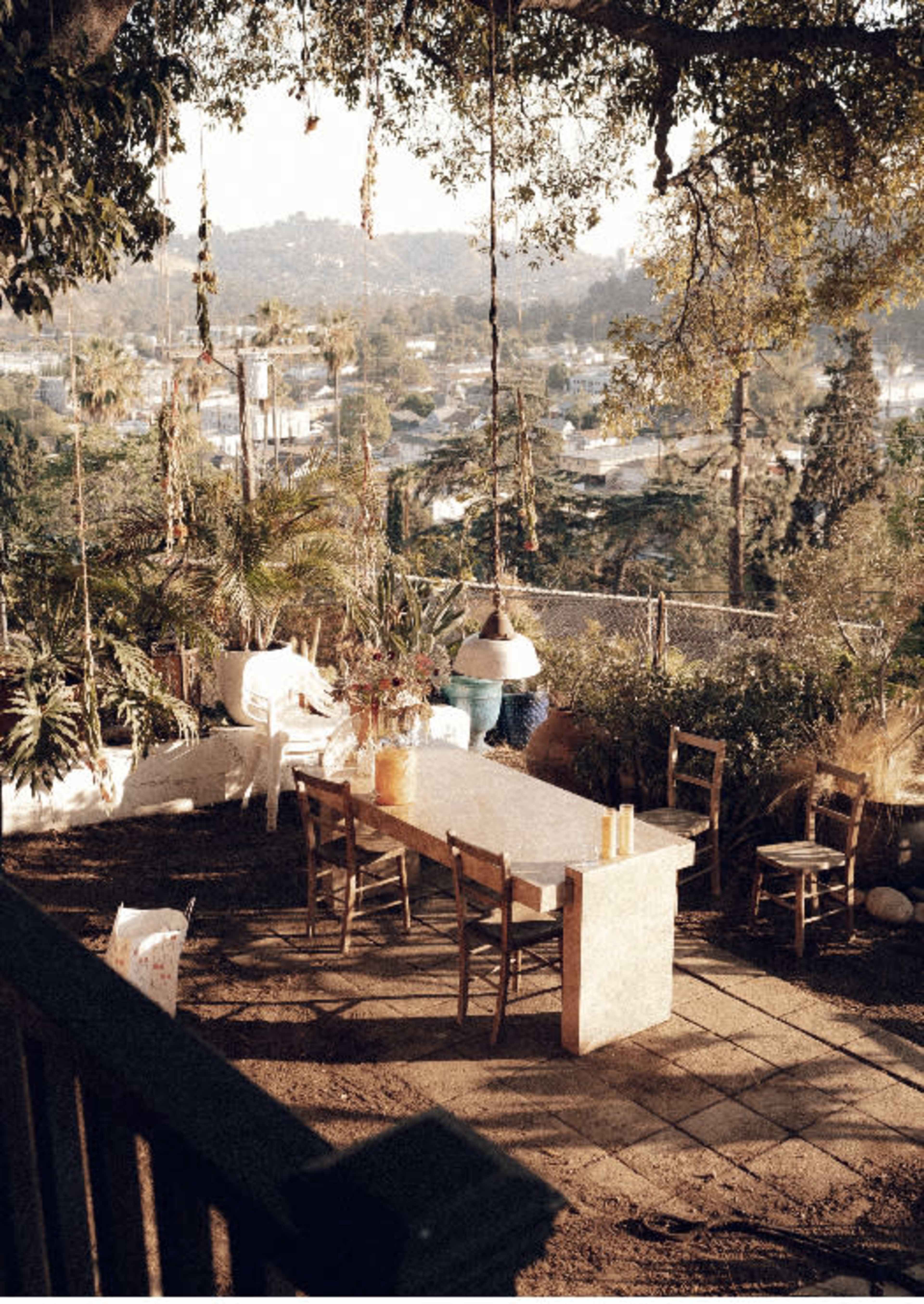 A long stone table surrounded by wooden chairs is set in a garden with hanging plants and a view of distant hills.