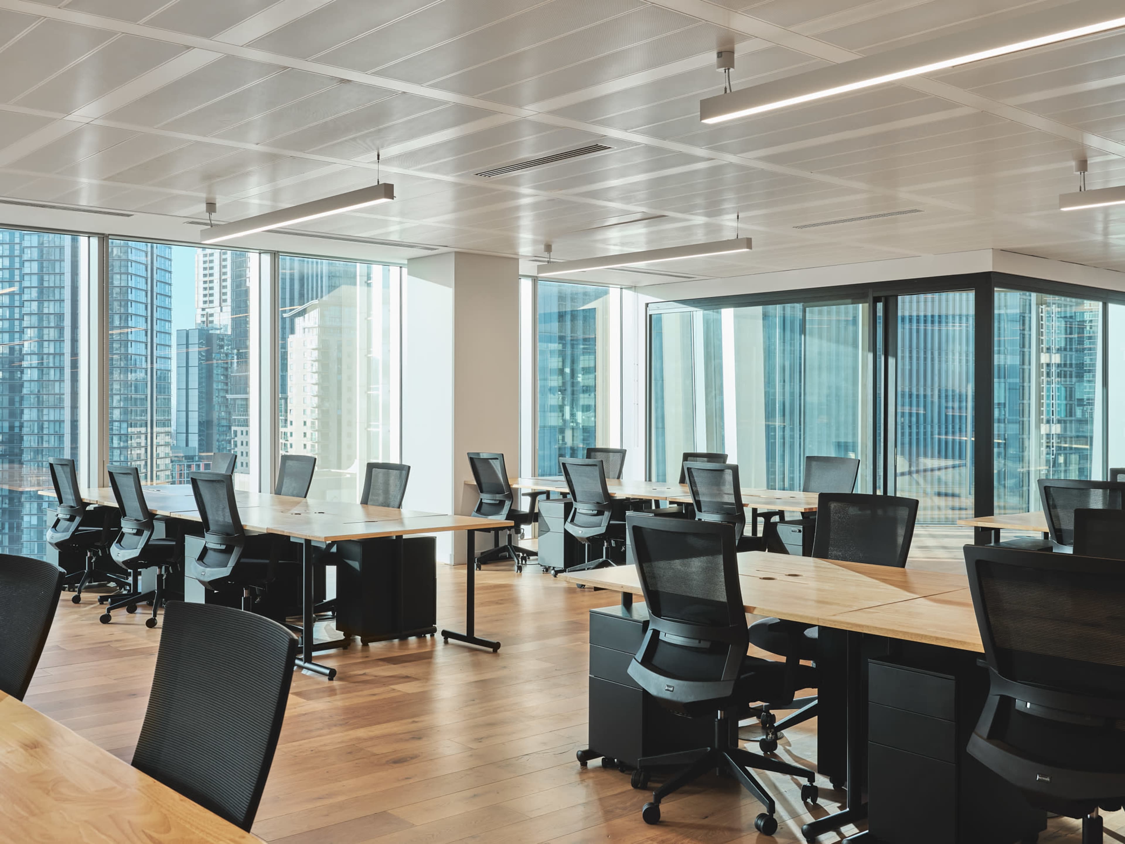 A modern office space with several workstations featuring black chairs and wooden desks, surrounded by large windows that provide a view of a city skyline.