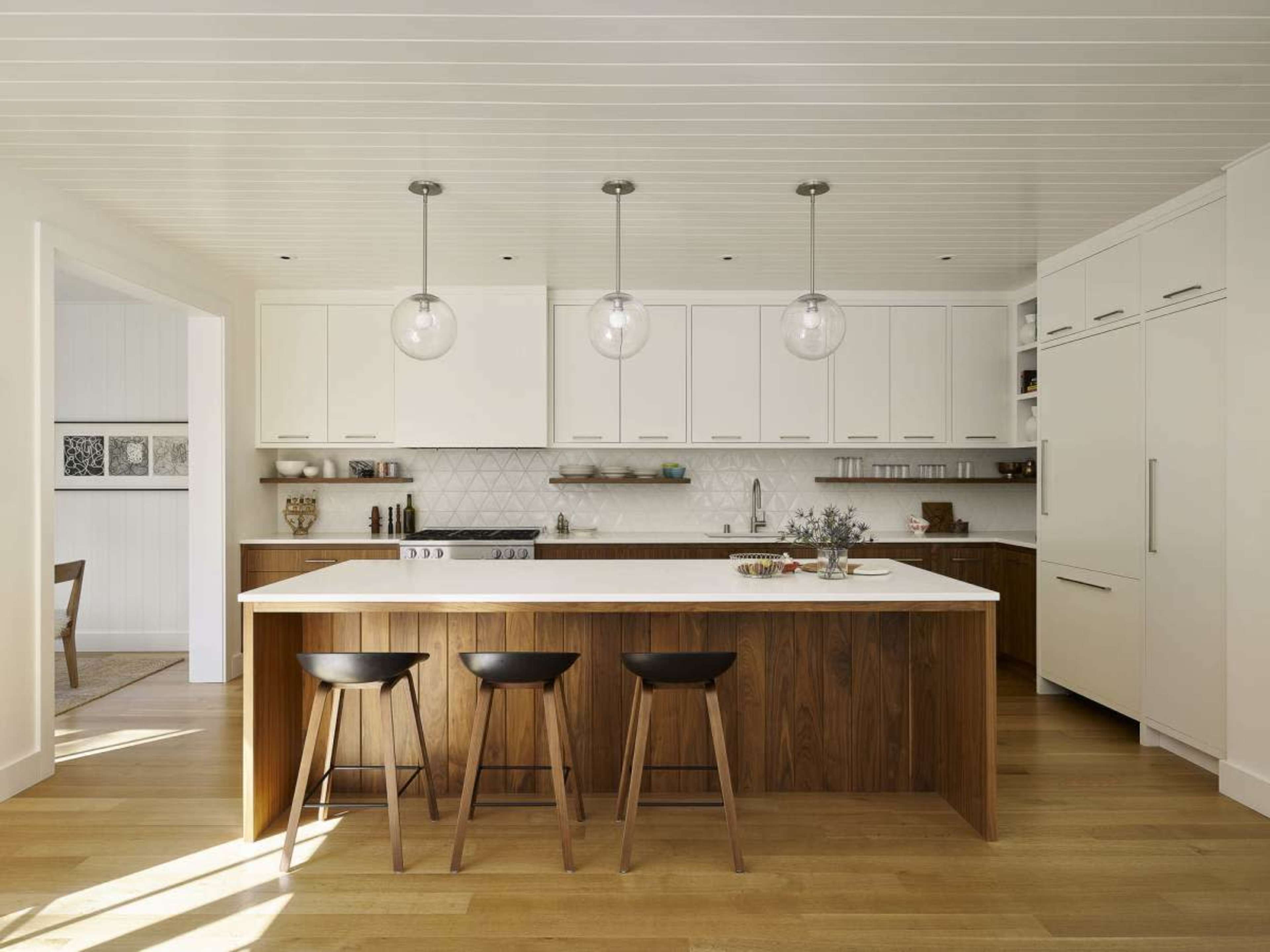 A modern kitchen features a central island with three wooden stools, white cabinetry, and pendant lighting.