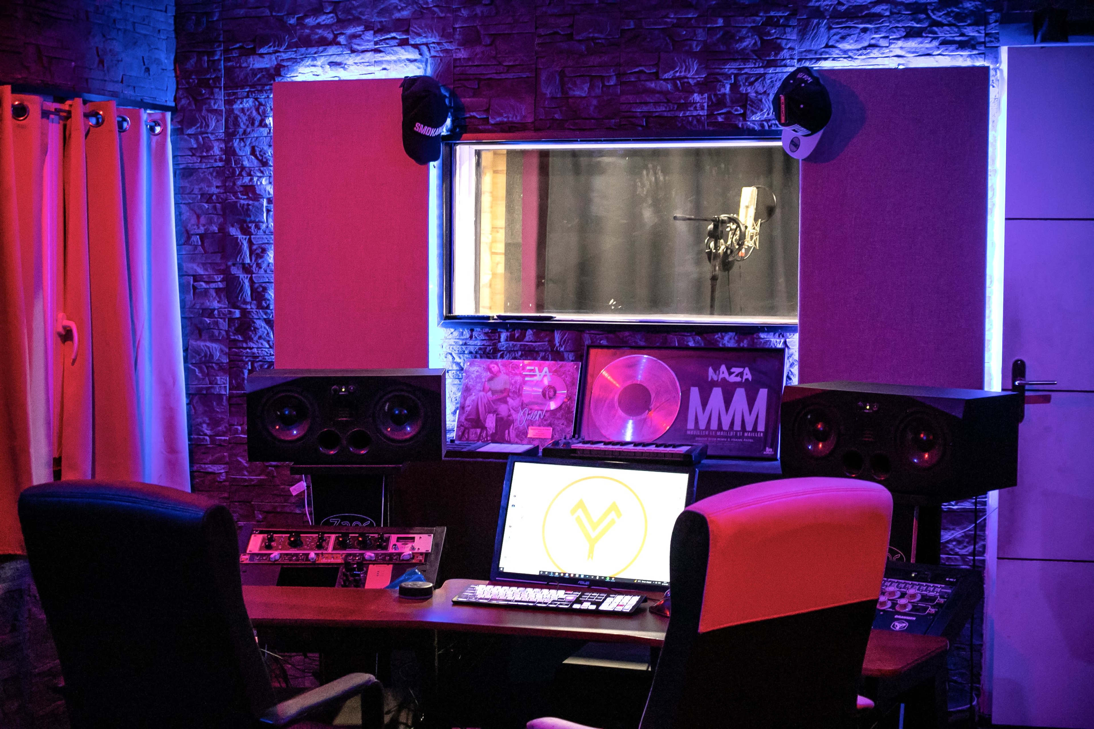 A modern music studio with a microphone in a soundproof booth, large speakers, and a computer setup on a wooden desk.