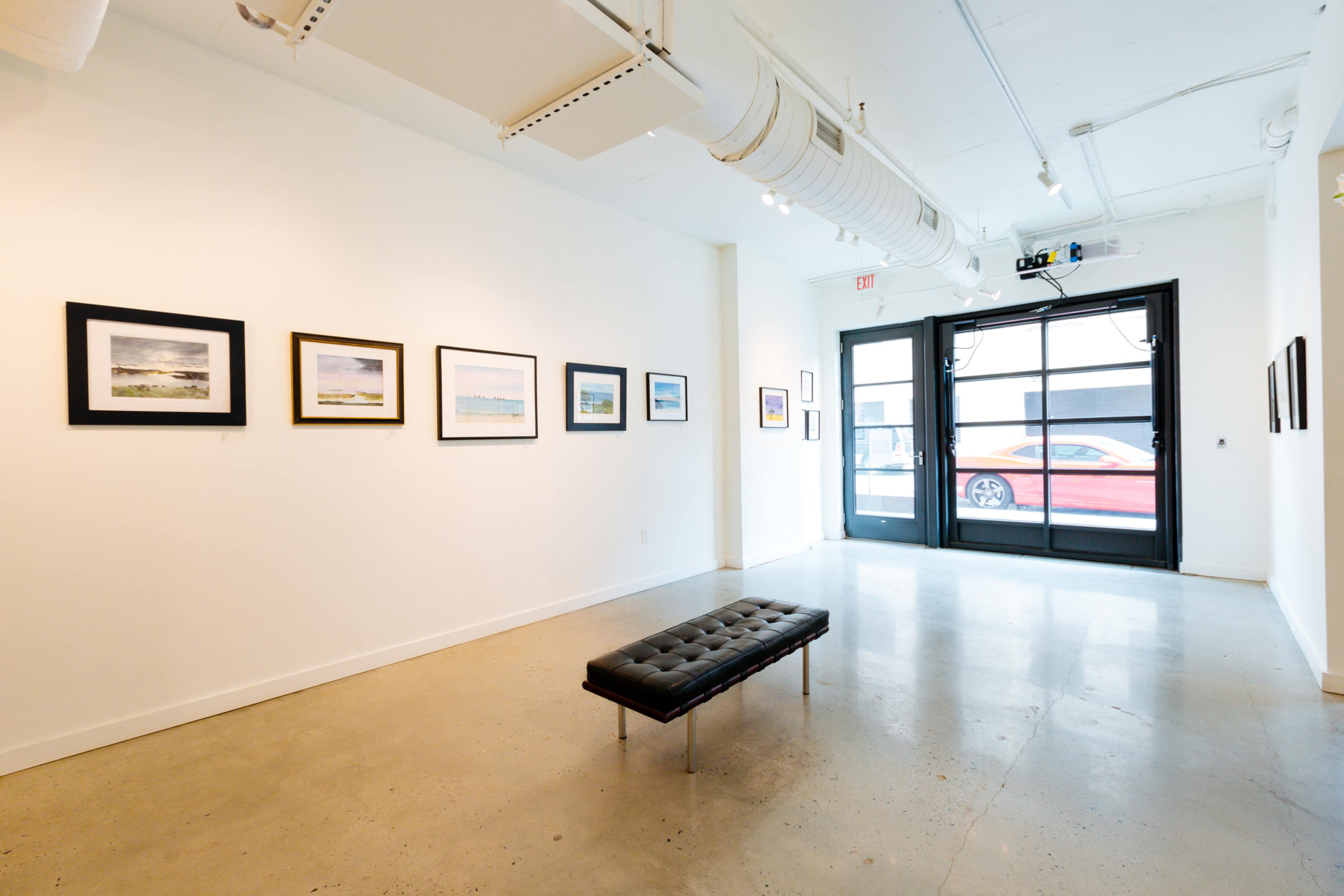 The image shows a gallery interior with framed photographs on white walls, a black bench in the center, and large windows that allow light in.