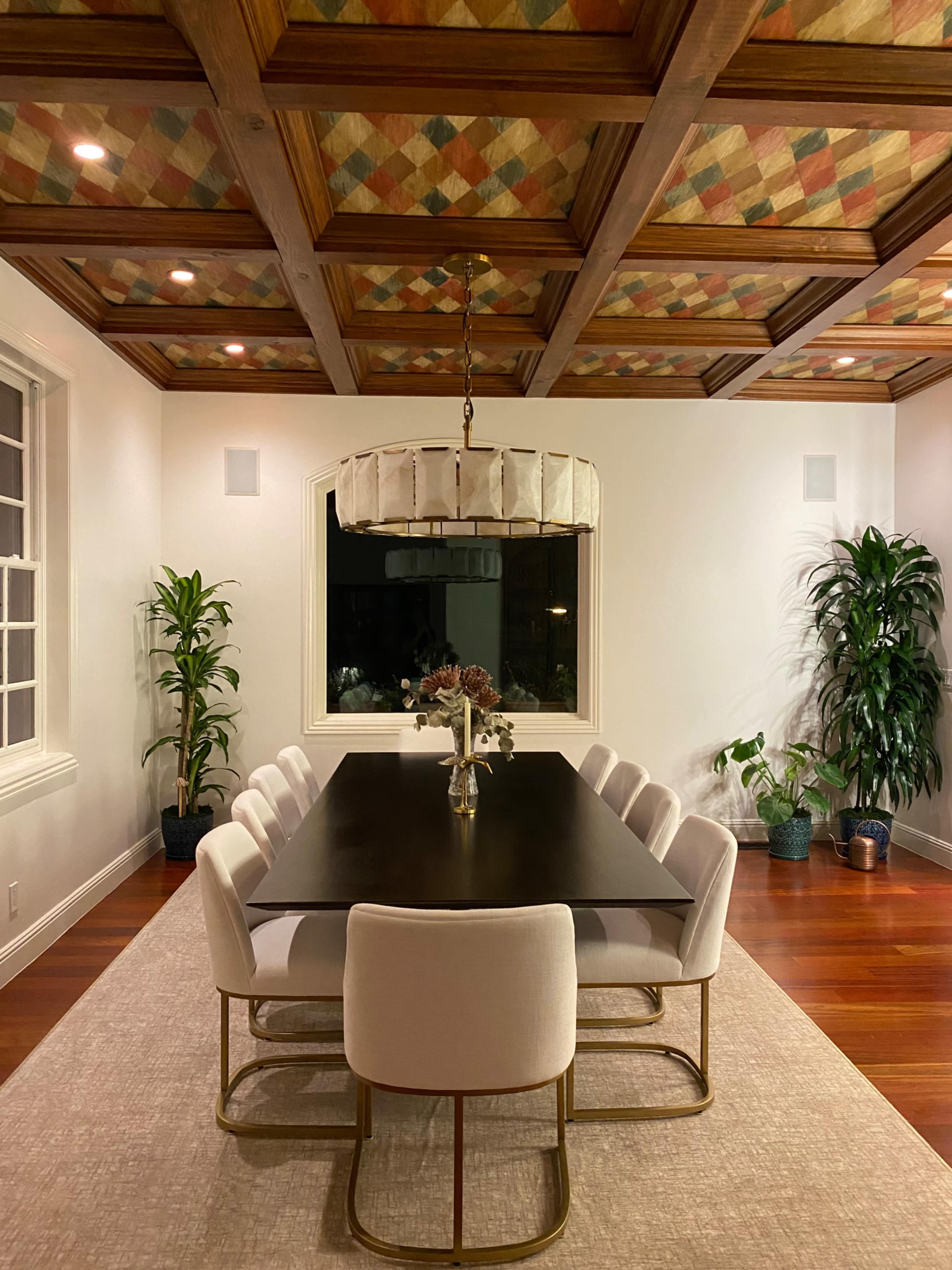 A modern dining room features a long dark table surrounded by light-colored upholstered chairs beneath a patterned ceiling and a decorative light fixture.