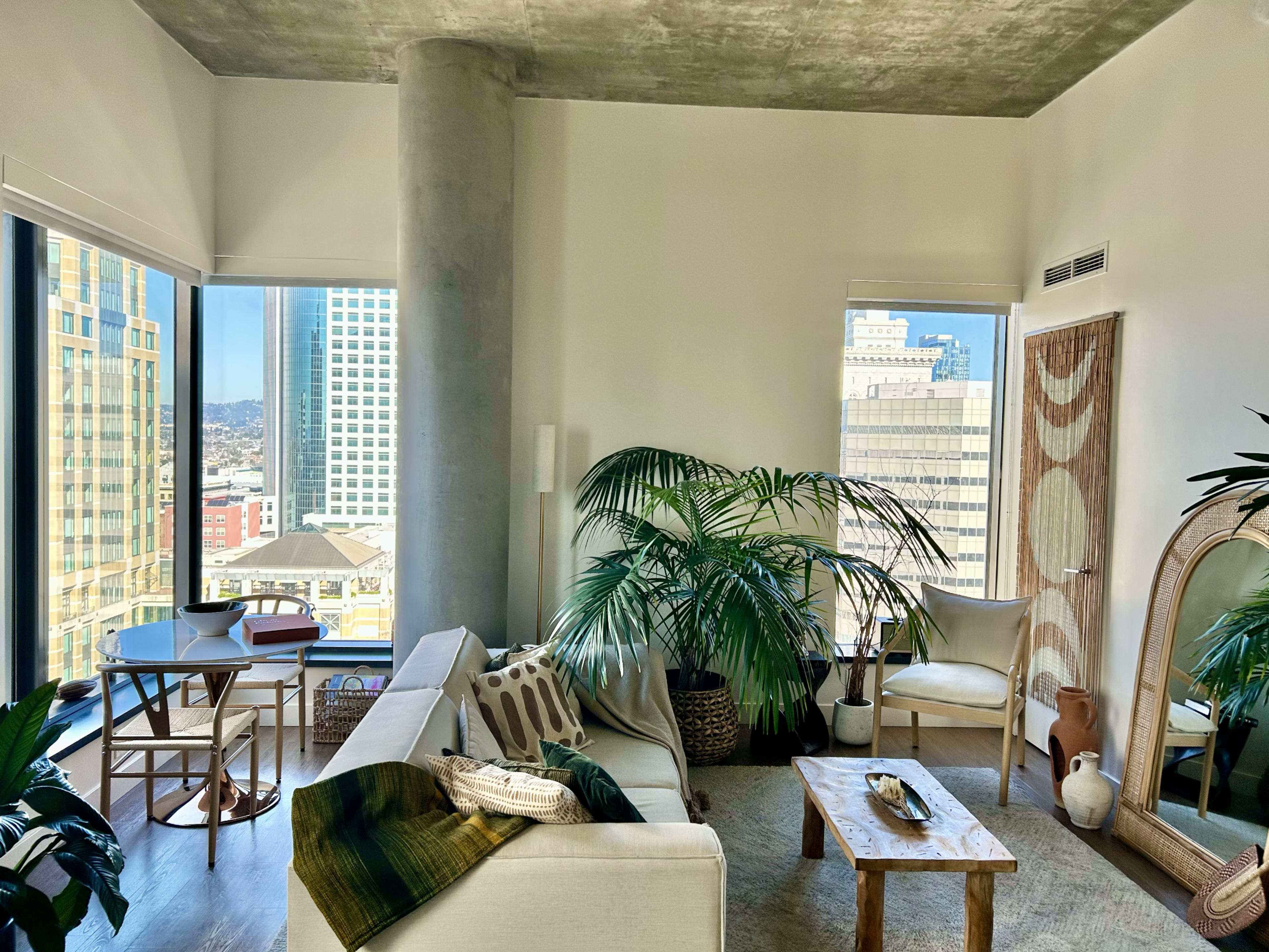 The image shows a modern living room featuring a light-colored sofa, potted plants, a wooden coffee table, and a view of city buildings through large windows.