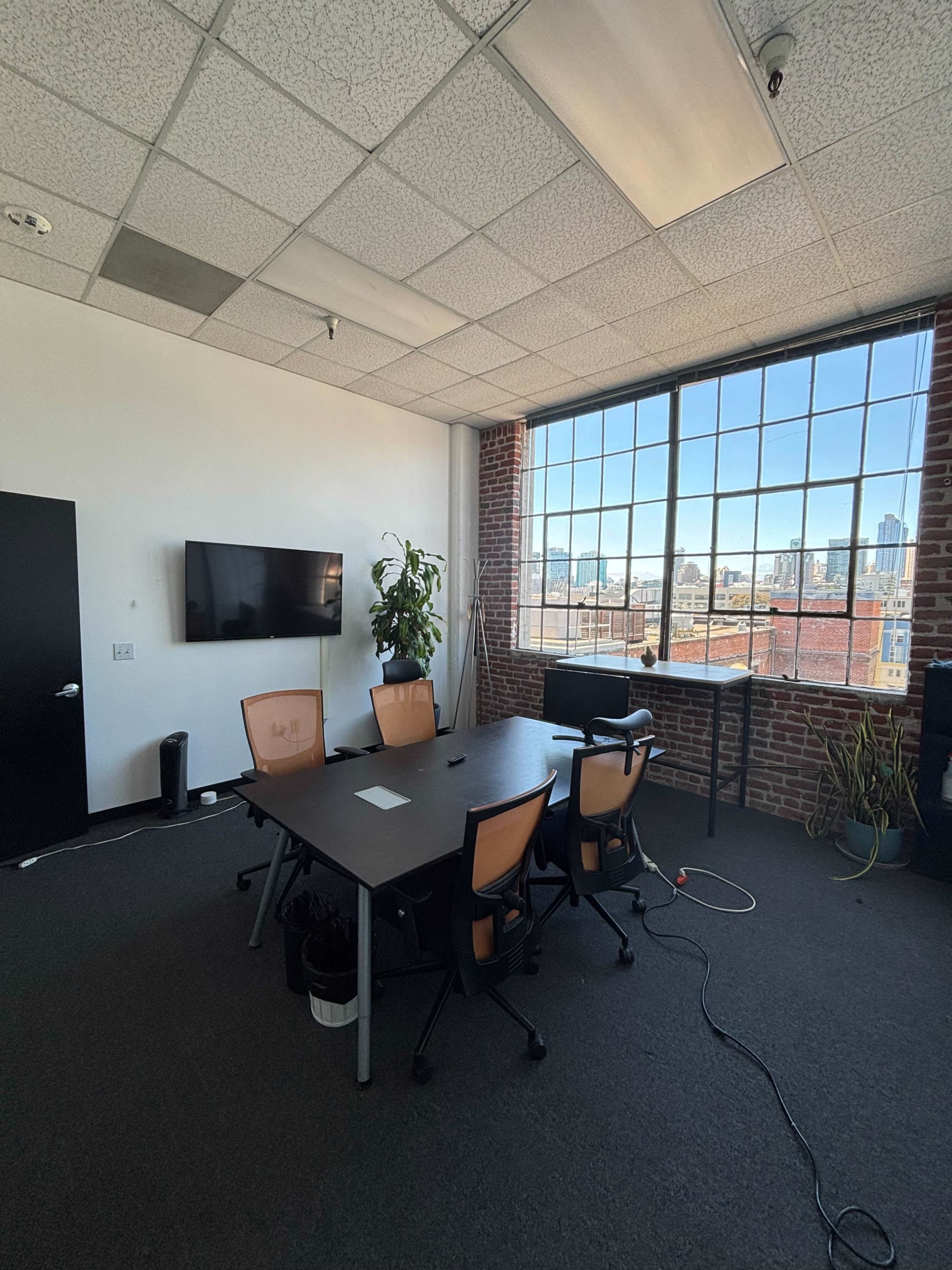 The image shows a modern conference room with a large table, several chairs, a TV, and a view of a city skyline through large windows.