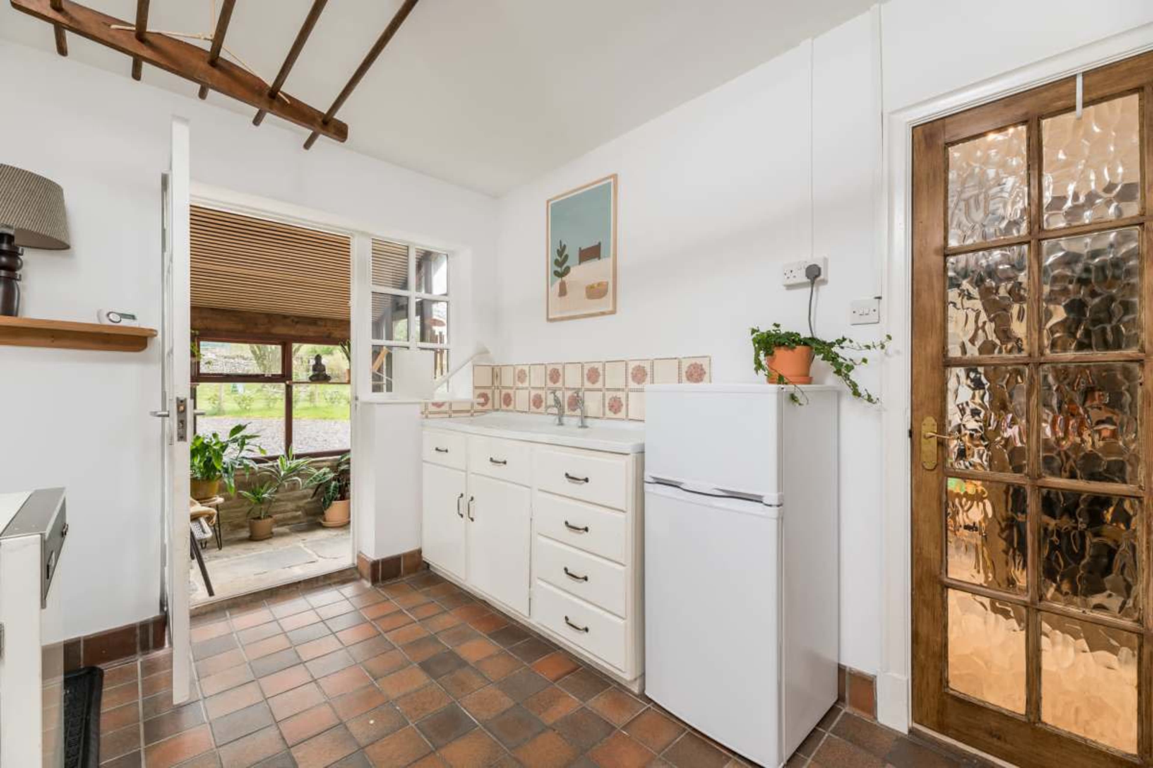 The image shows a kitchen area with a white refrigerator, sink, and a door leading to an outdoor space with plants.