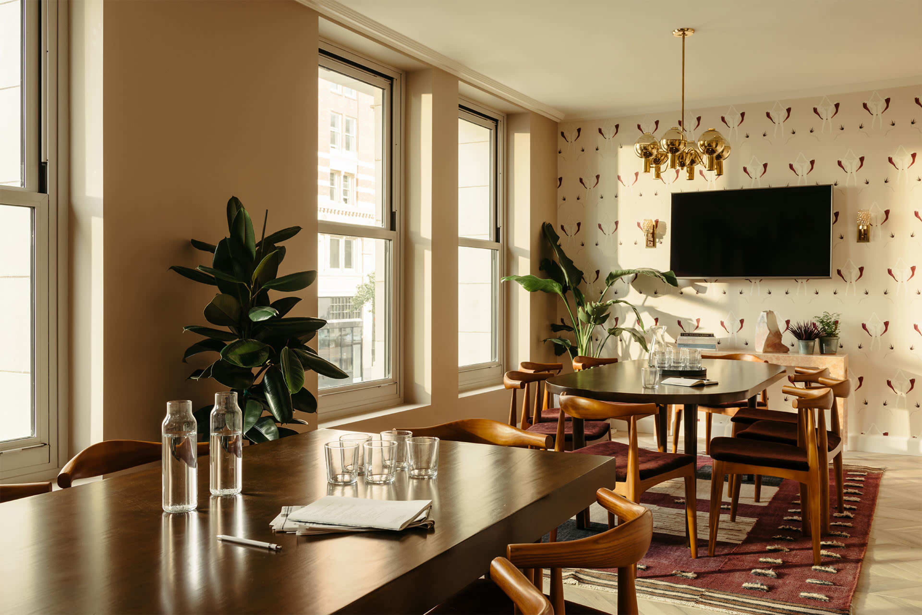 A modern conference room features a long wooden table, chairs, plants, large windows, and a wall-mounted screen.