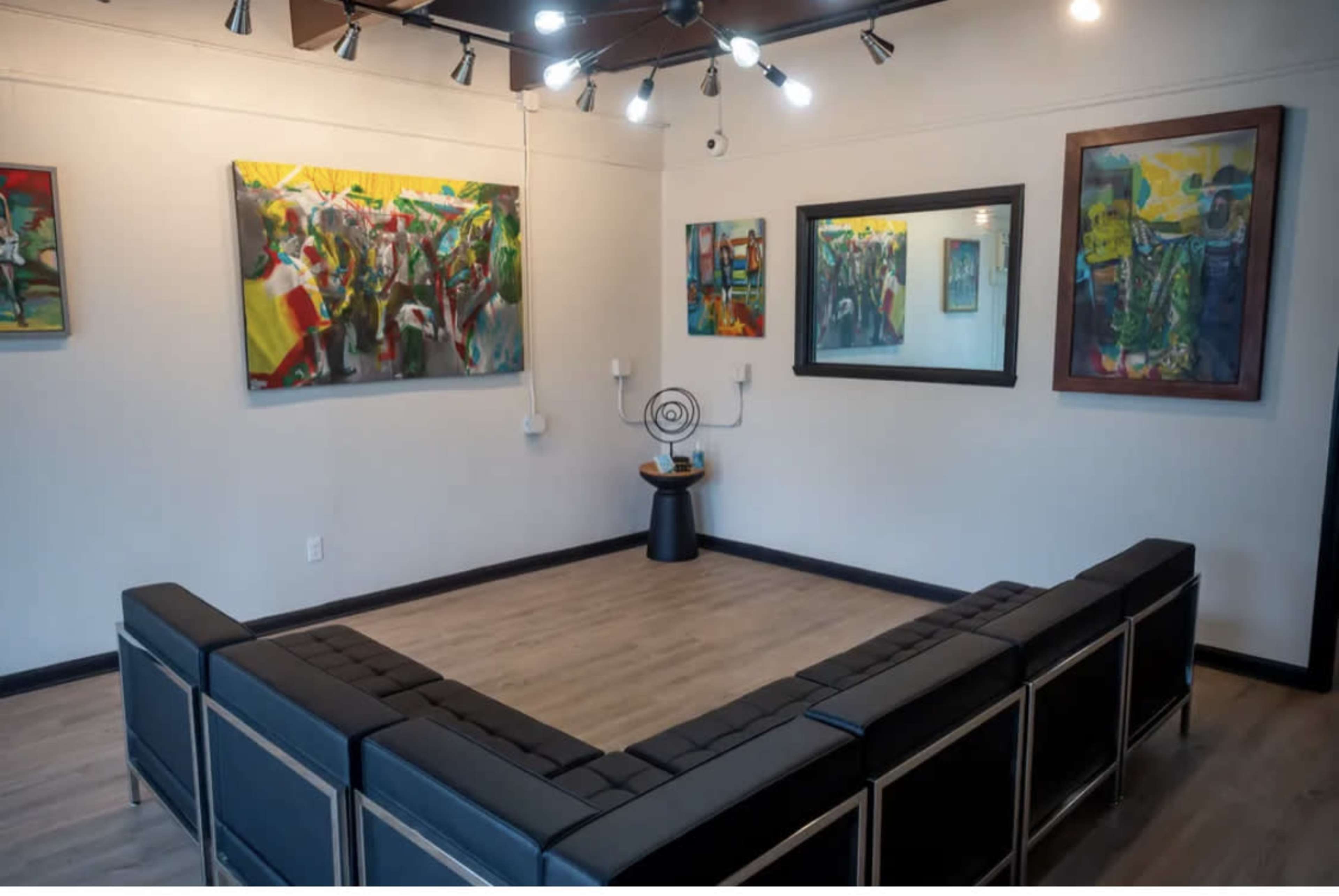 The image shows a modern art gallery with a U-shaped seating arrangement facing colorful paintings on the walls and a round decorative piece in the center.