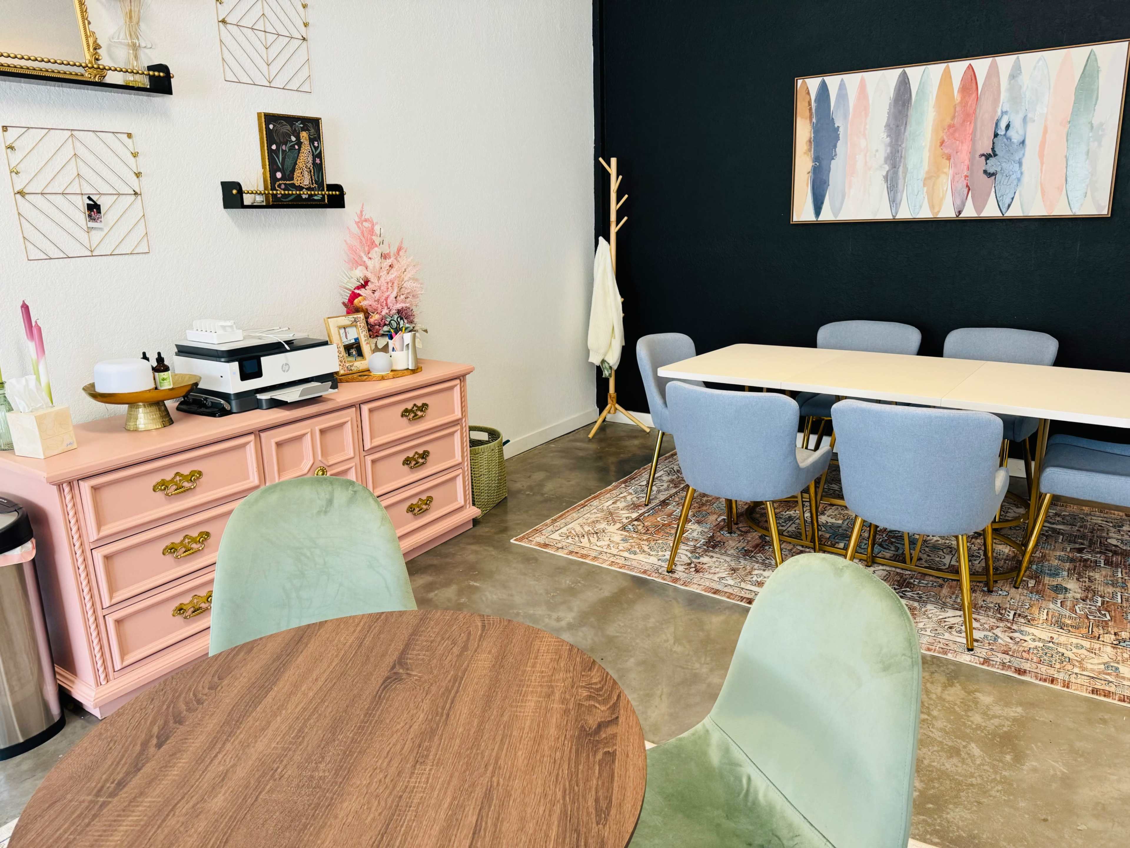 A modern office space featuring a pink sideboard, a round wooden table, grey chairs, and a black accent wall adorned with decorative artwork.