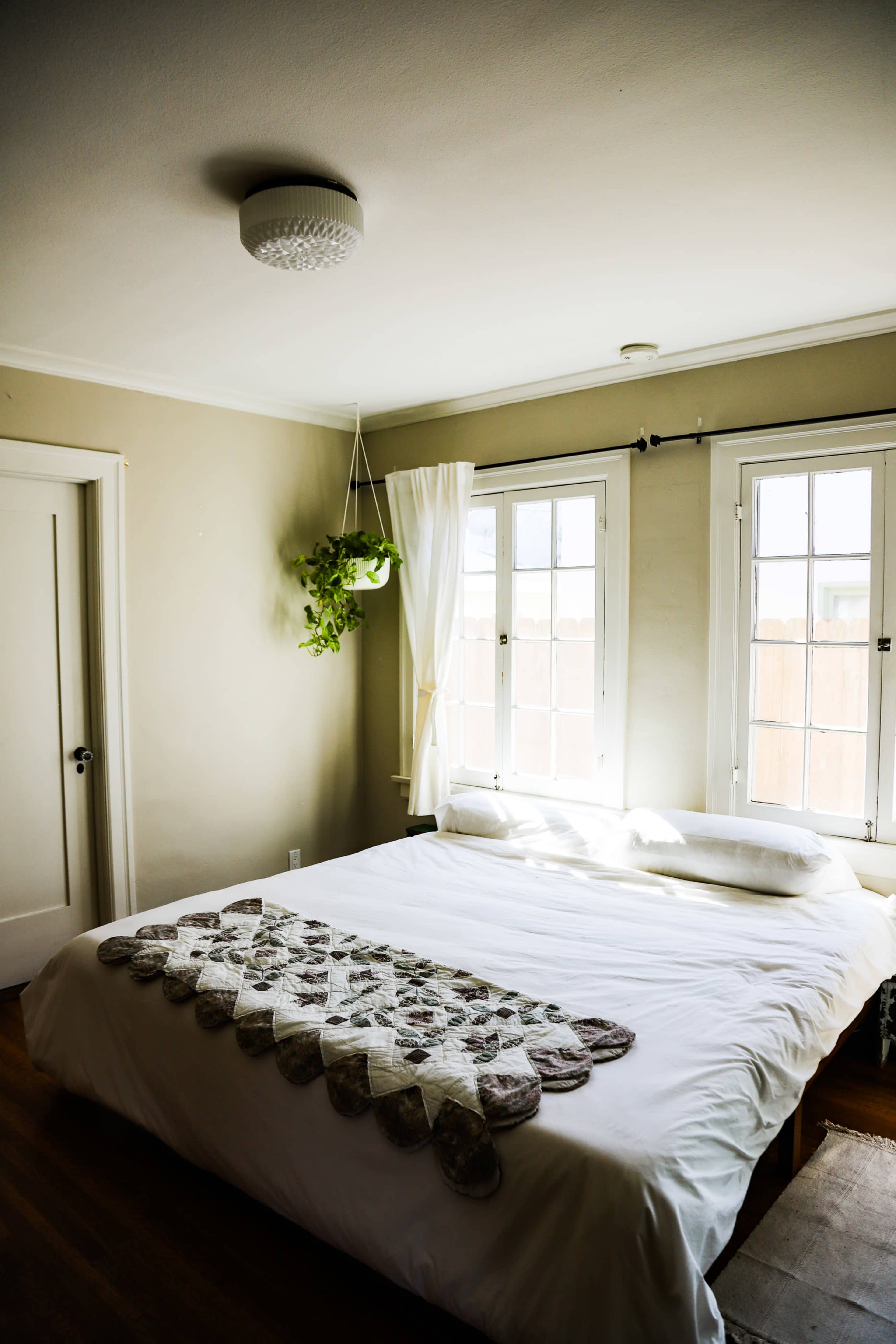 A minimally furnished bedroom features a bed with white linens and a decorative mat, illuminated by natural light through two windows.