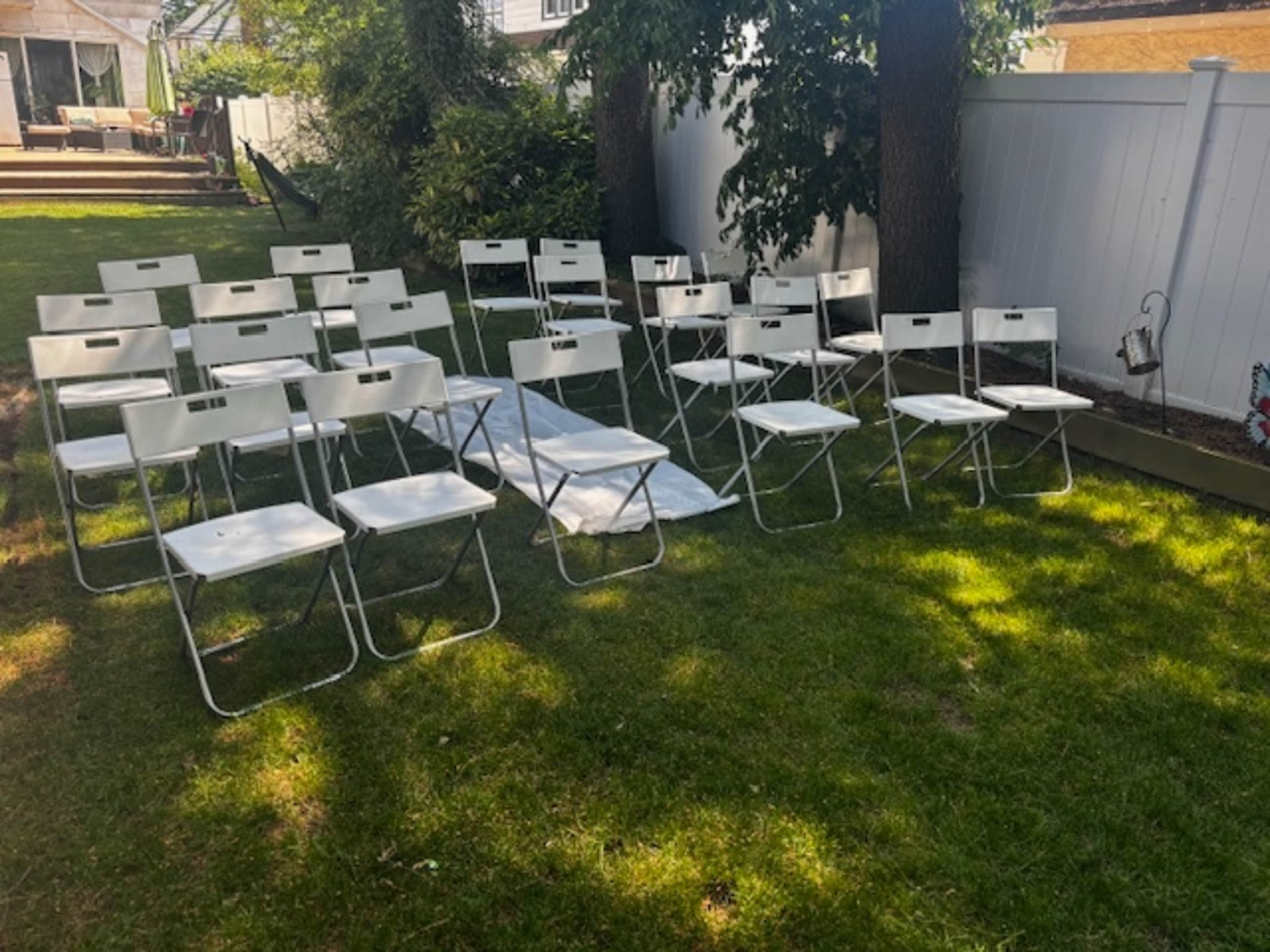 A set of white folding chairs is arranged in rows on a grassy area under trees.