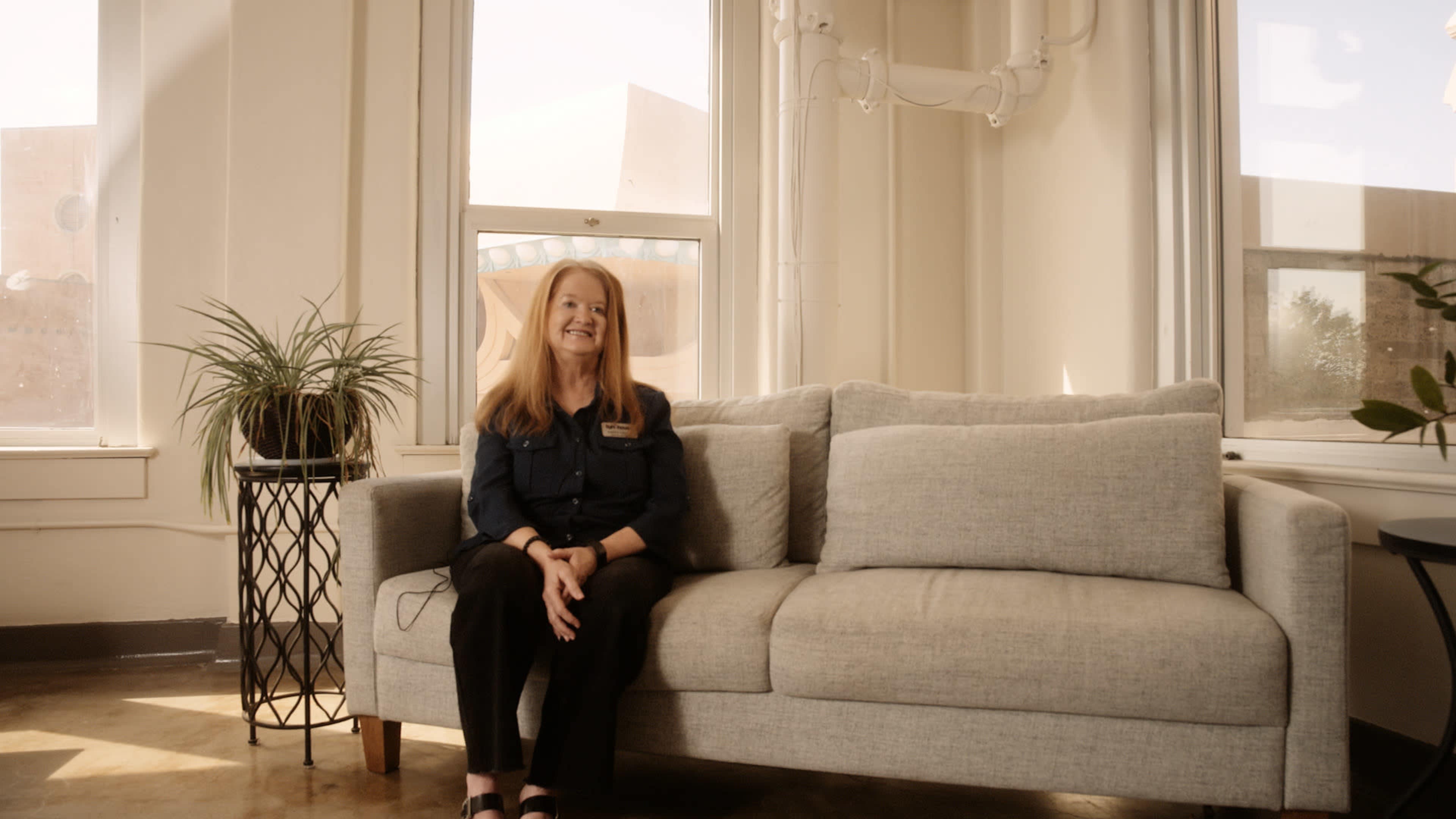 A woman sits on a gray couch in a well-lit room with windows and a potted plant nearby.