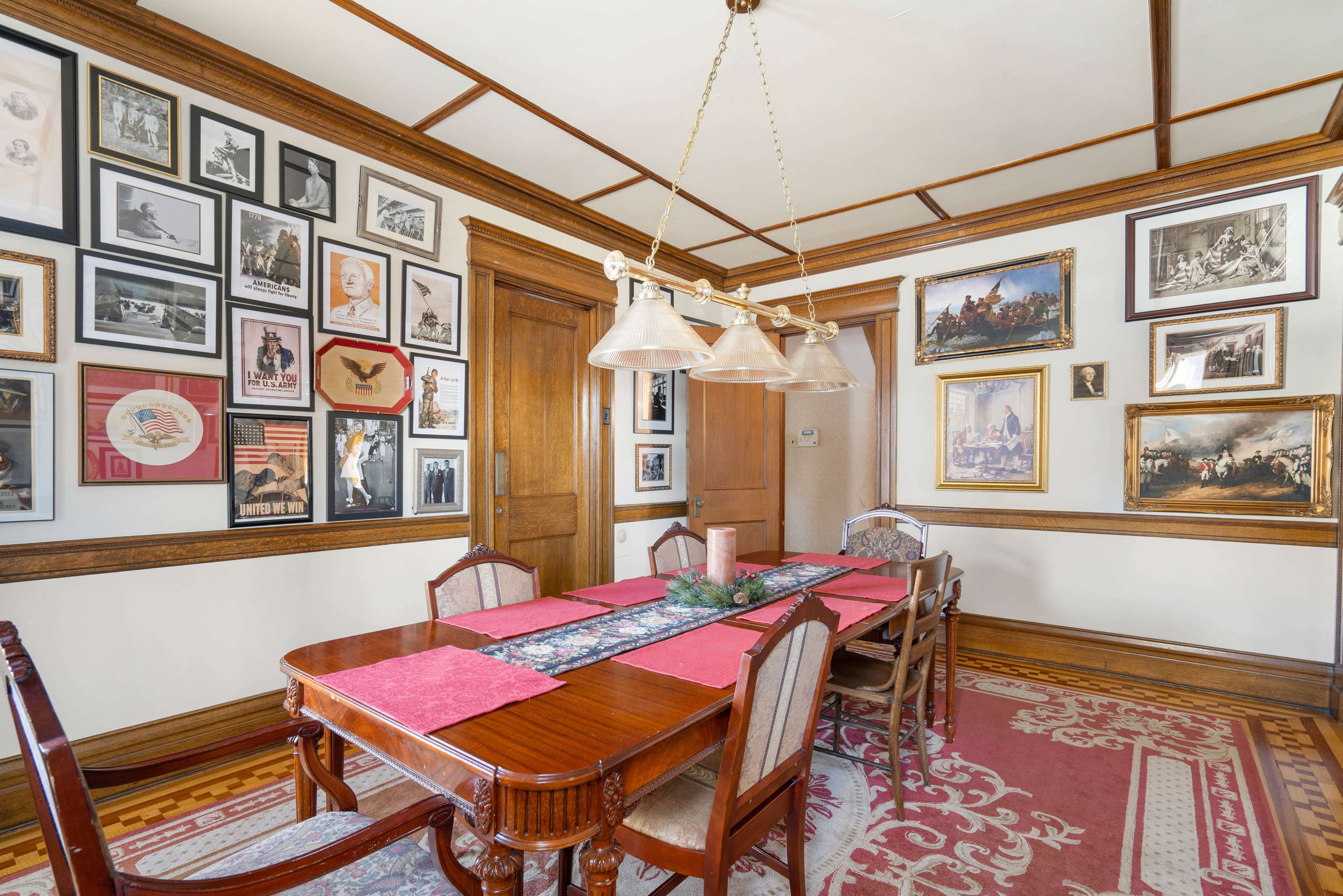 The image shows a dining room featuring a wooden table with red placemats, surrounded by chairs, and walls adorned with framed photographs and paintings.