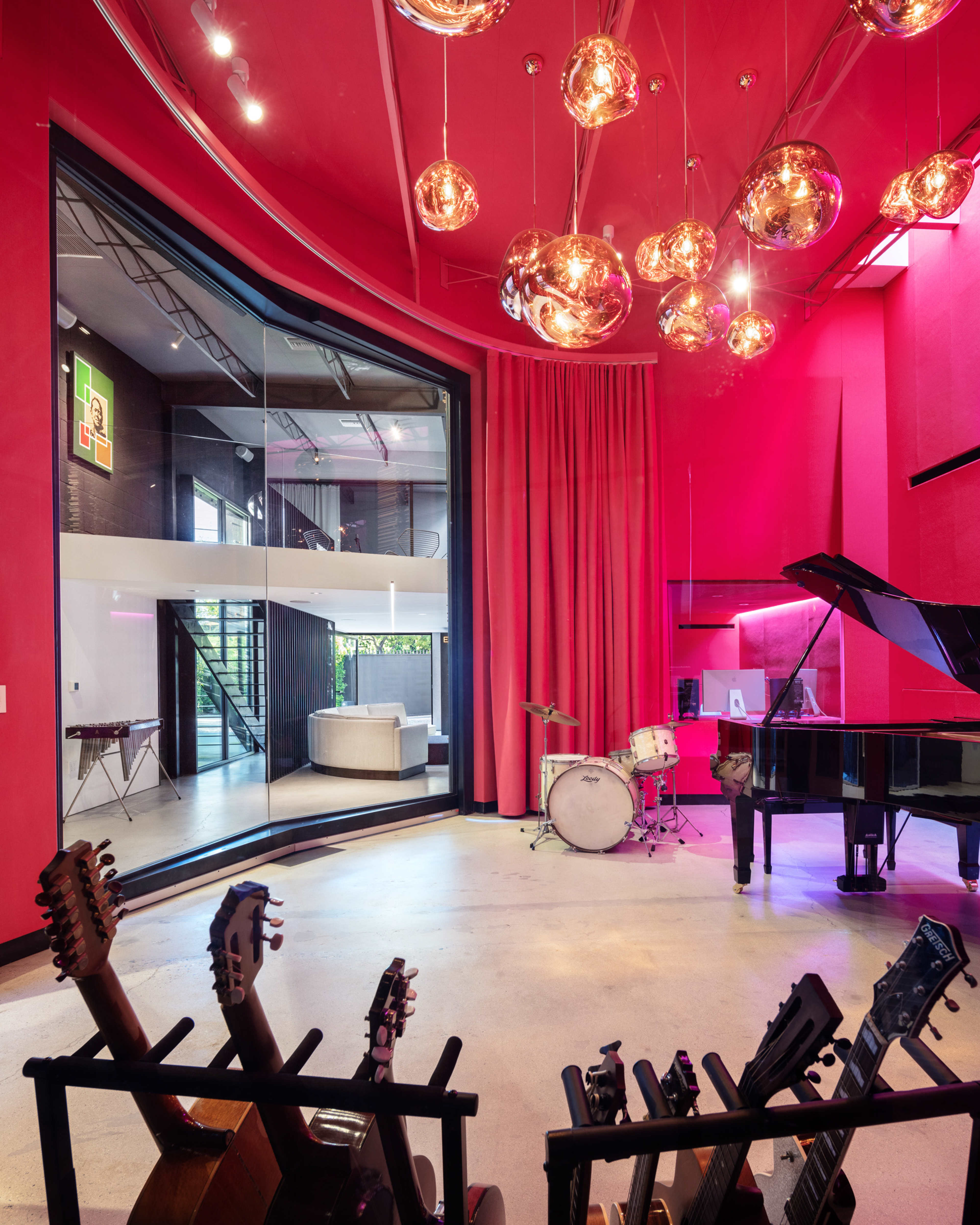 A vibrant music studio features a grand piano, drums, and several guitars displayed on stands, with large windows providing a view of an adjacent living space.