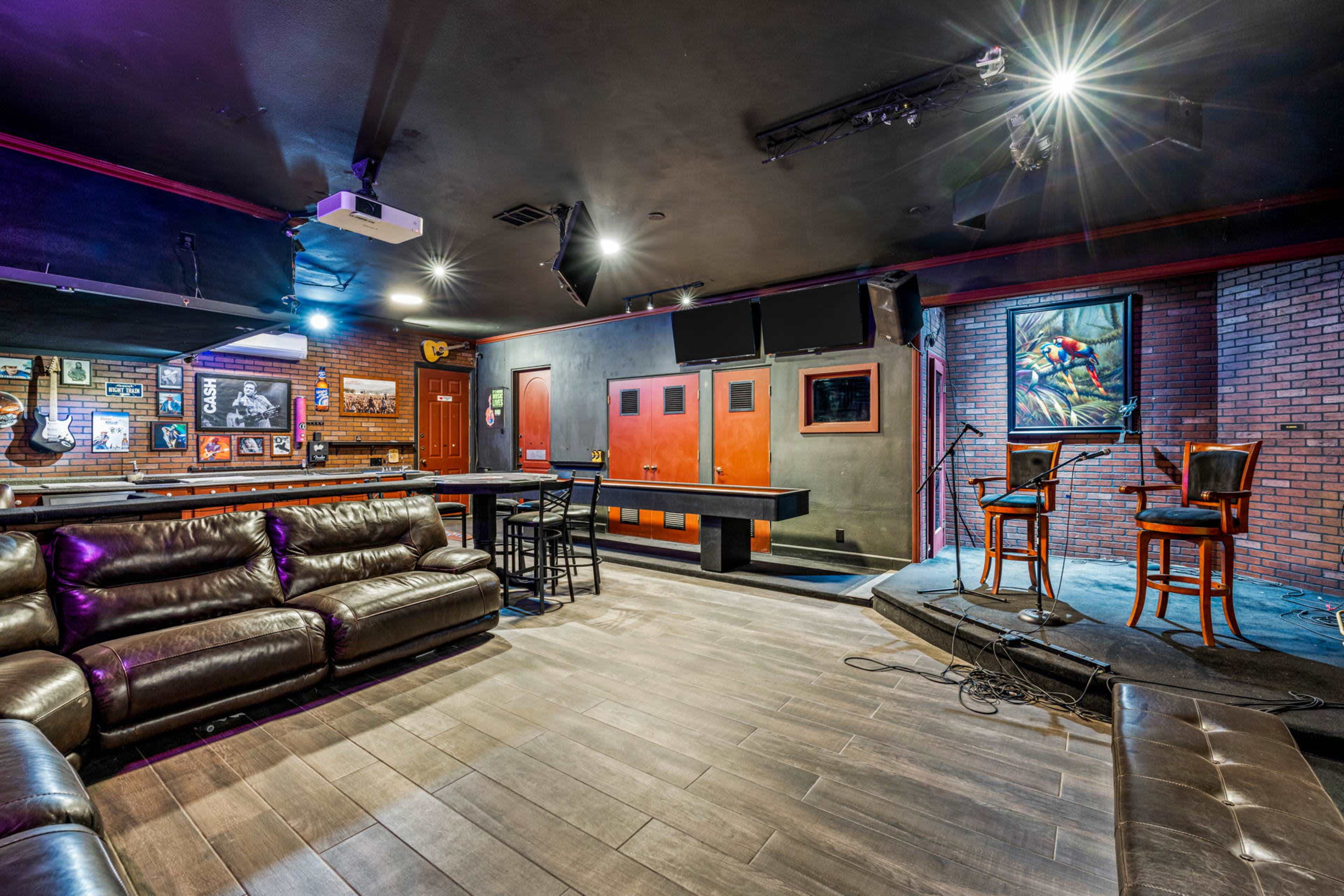 The image shows an indoor performance space featuring a small stage with two chairs, a bar area, and lounge seating arranged in a room with wooden flooring and a brick wall.