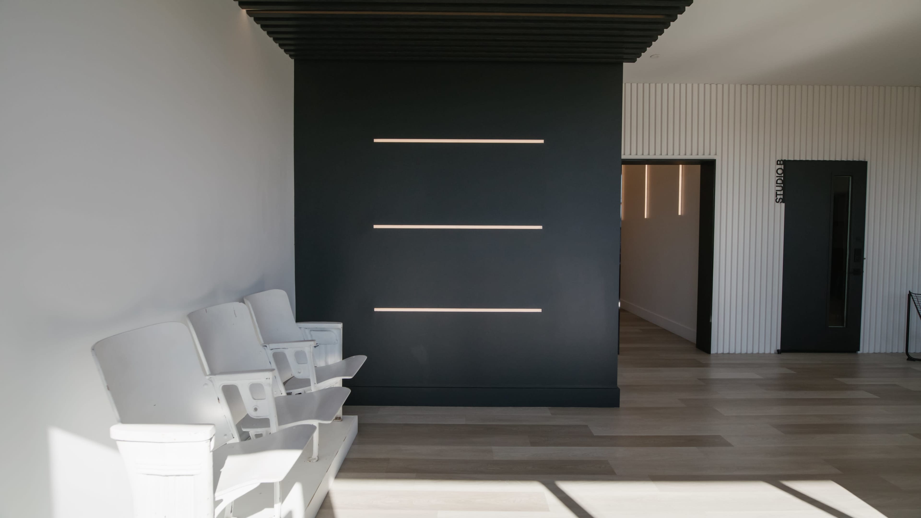 The image shows a minimalist interior space with a row of white chairs against a black wall featuring horizontal light strips, leading to an open doorway in the background.