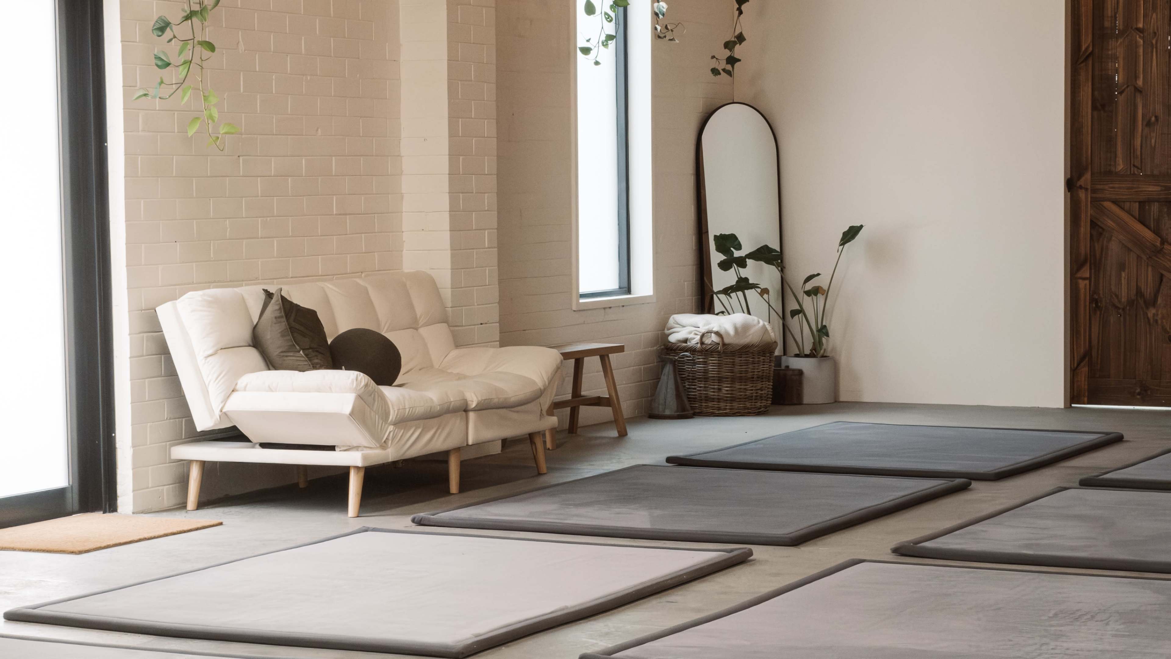 The room features a white couch, a mirror, and several gray mats arranged on the floor, with plants and a basket in the corner.