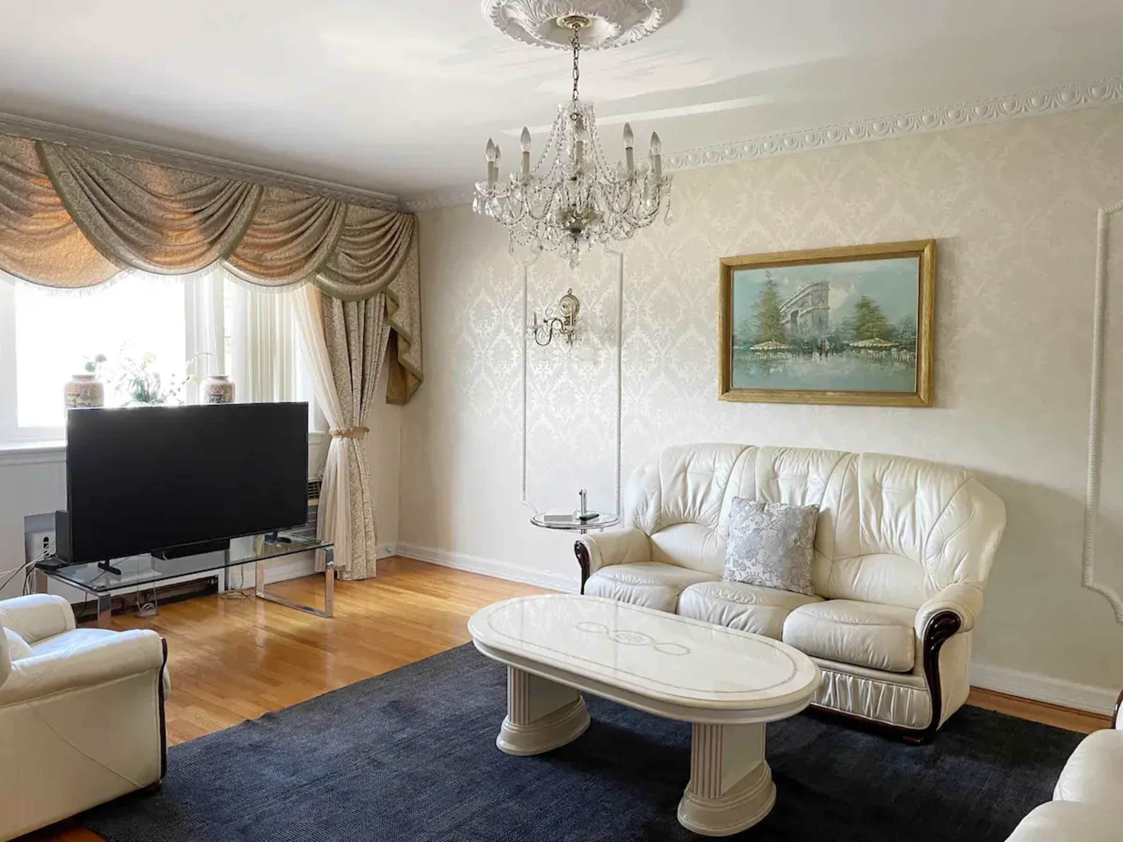 A living room features white leather sofas, a chandelier, a coffee table, and a large television against a patterned wall with an artwork above it.