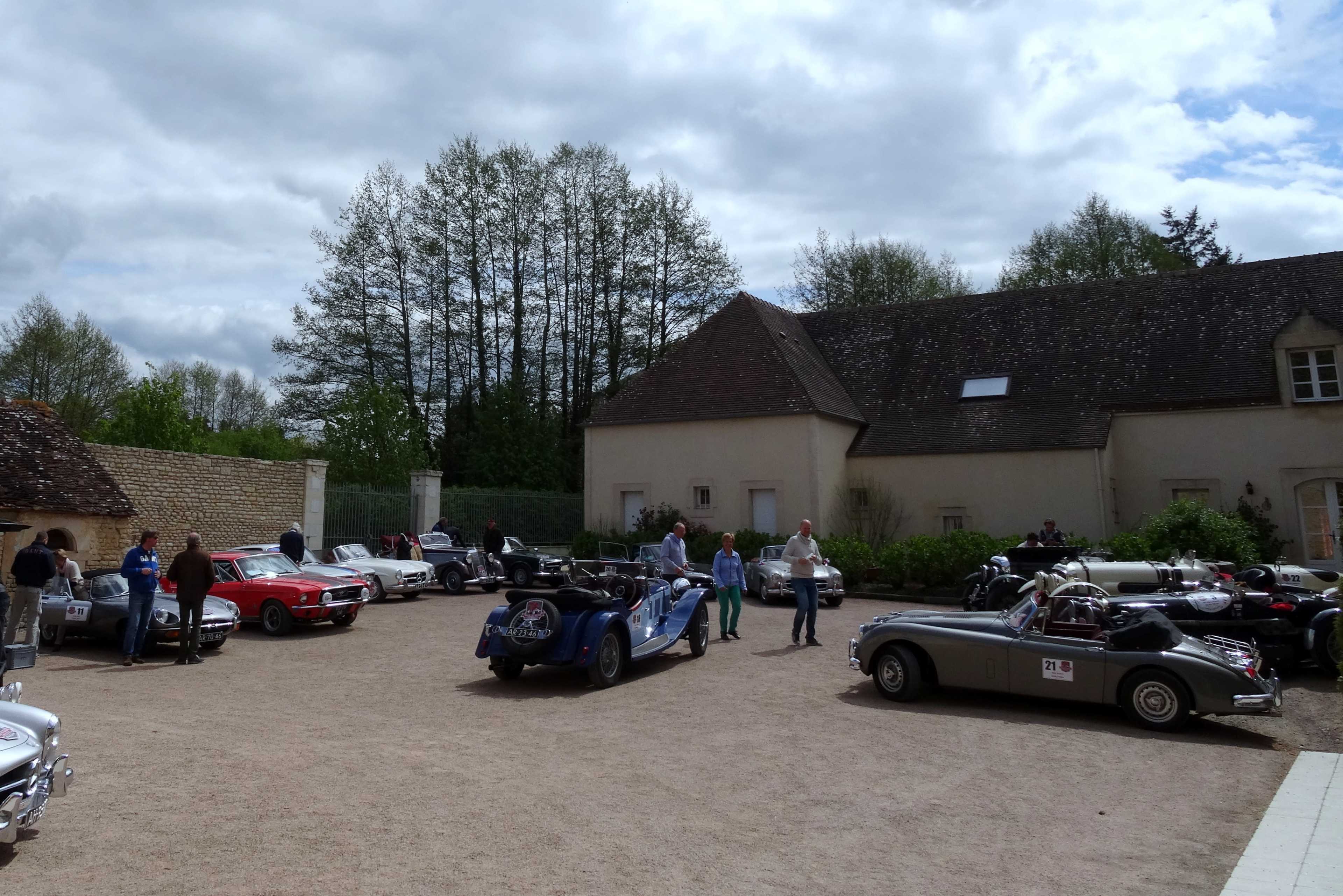 A gathering of vintage cars is set in a courtyard next to a stone building surrounded by trees under a partly cloudy sky.