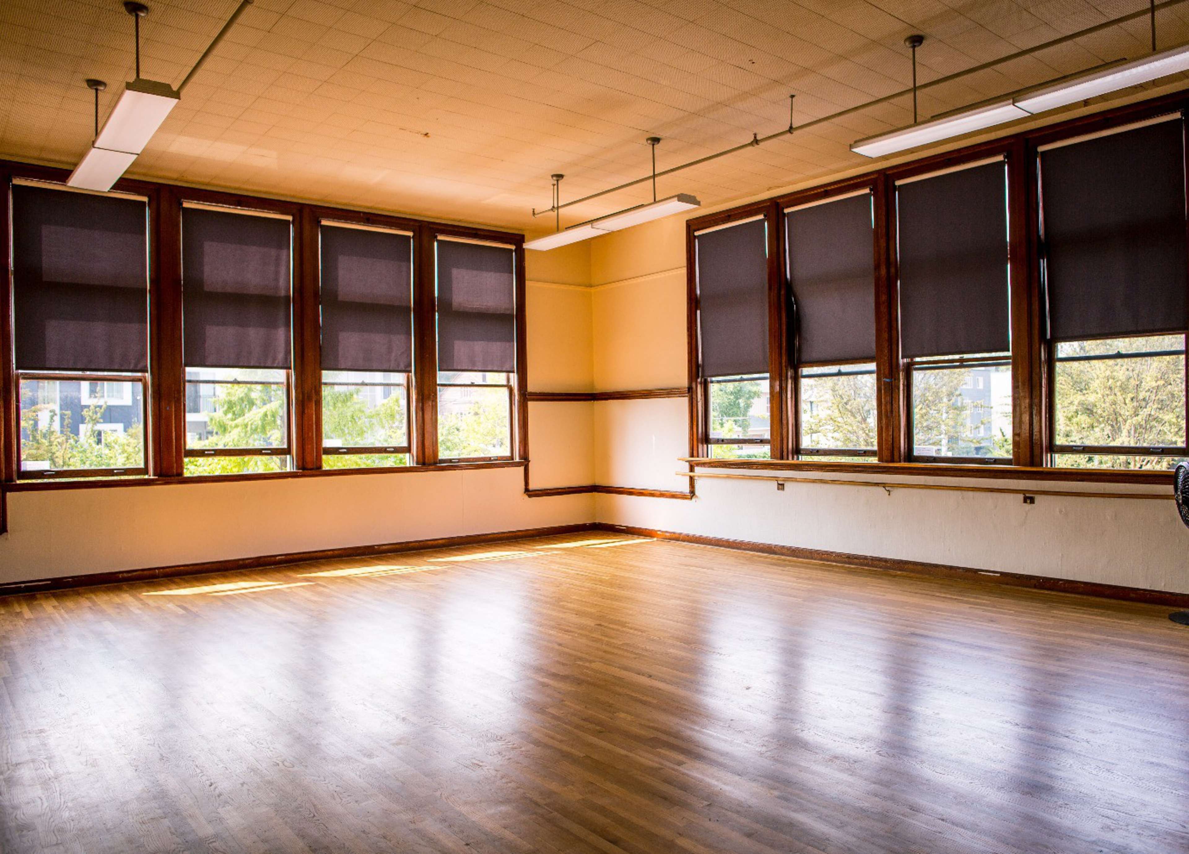 The image shows a spacious, empty classroom with large windows covered by blinds, allowing slivers of natural light to illuminate the wooden floor.