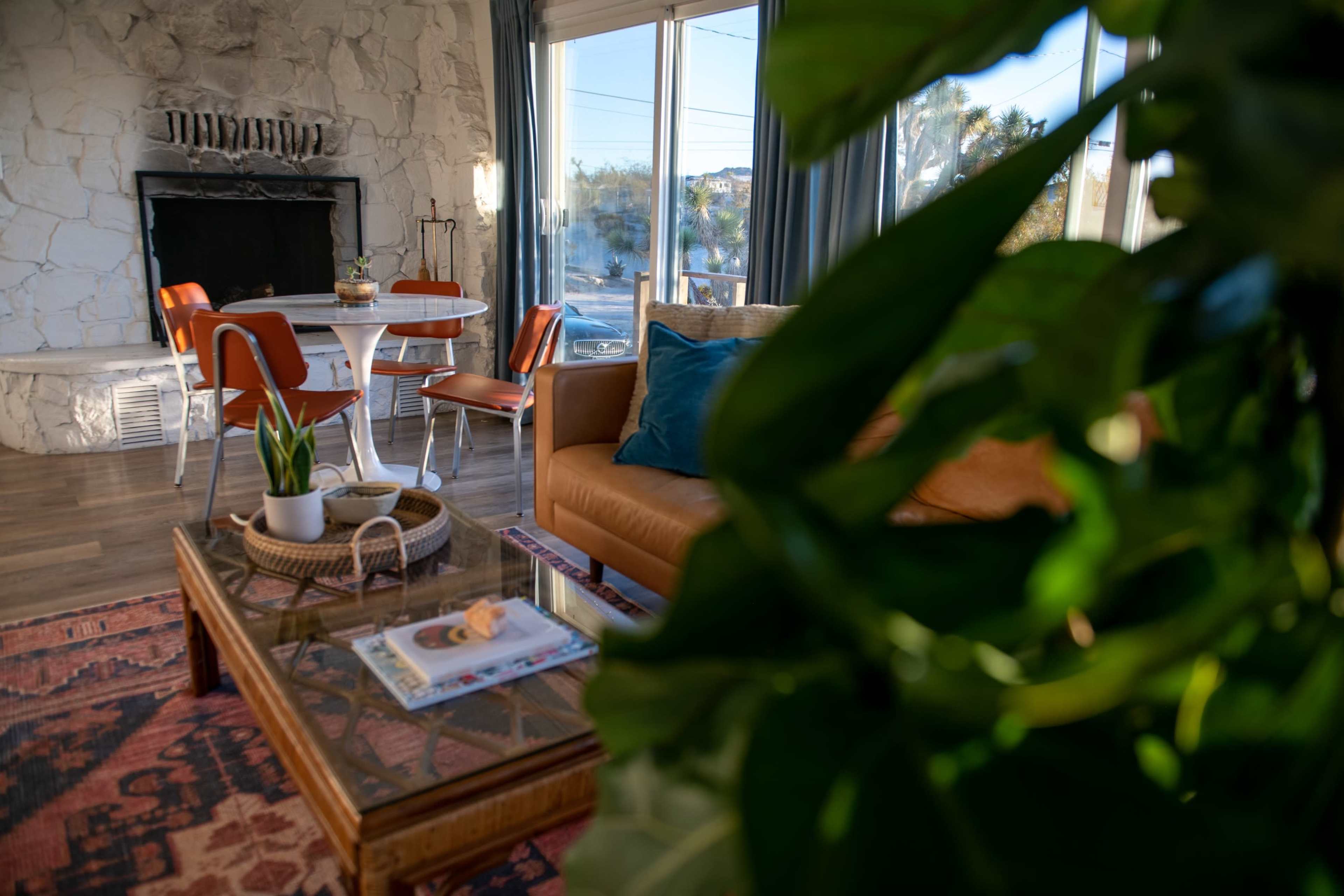 A cozy living room features a stone fireplace, a wooden coffee table with plants and magazines, and a dining area with a round table and orange chairs.