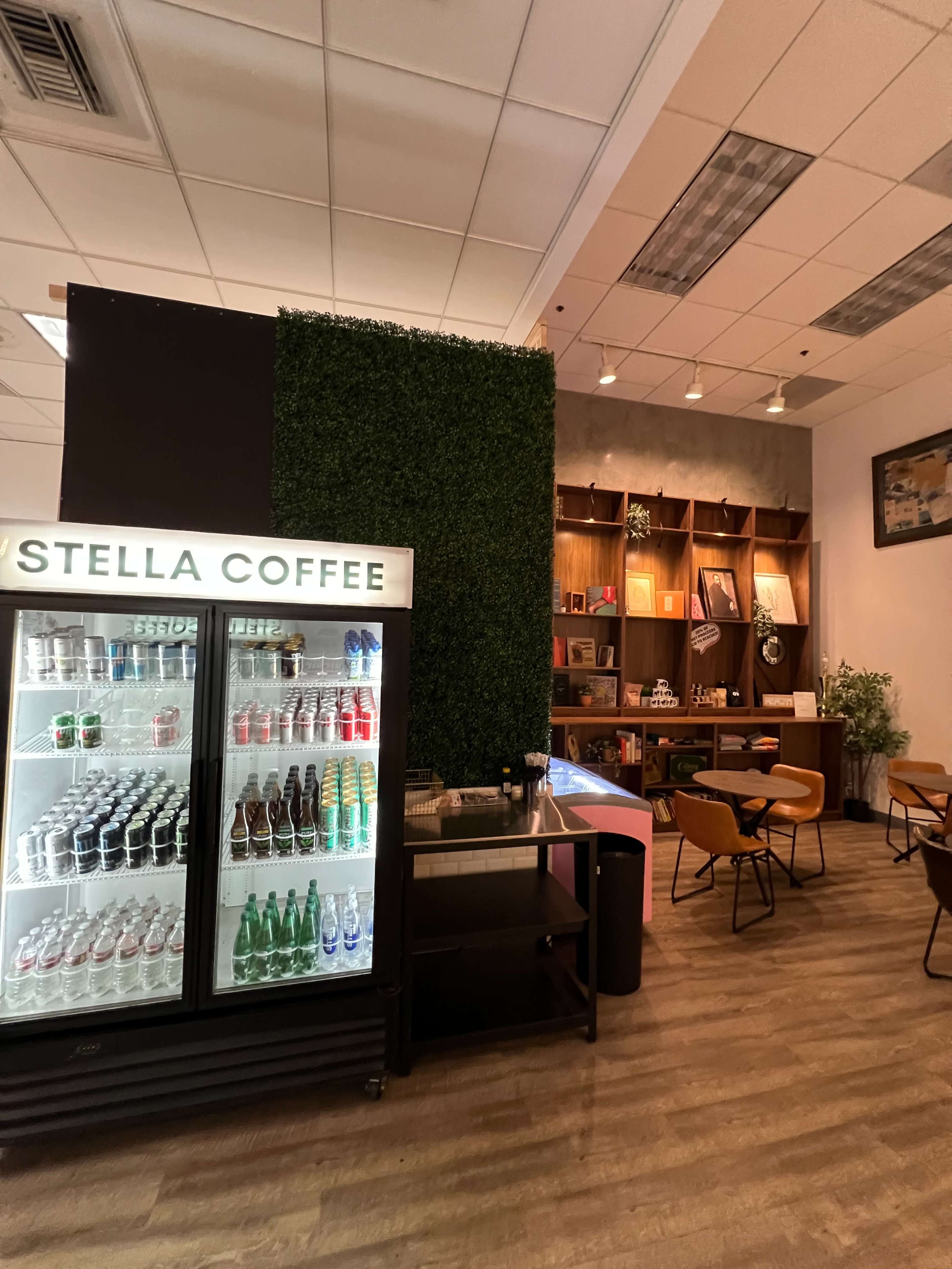 The image shows a coffee shop interior with a glass display refrigerator filled with beverages, a green wall, and wooden shelving containing books and decor.
