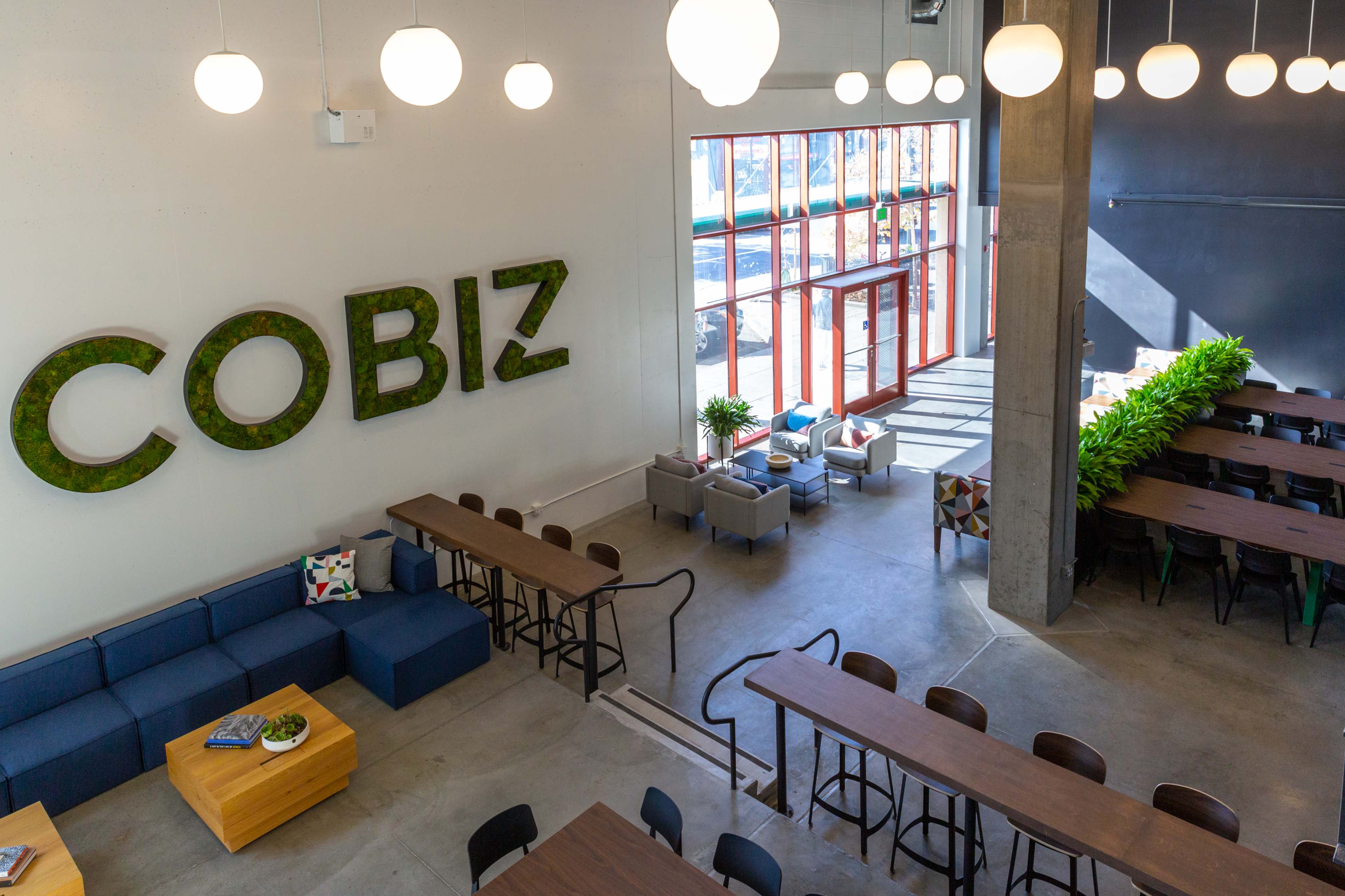 A spacious interior of a coworking space with large windows, green plants, and wooden tables arranged for seating.