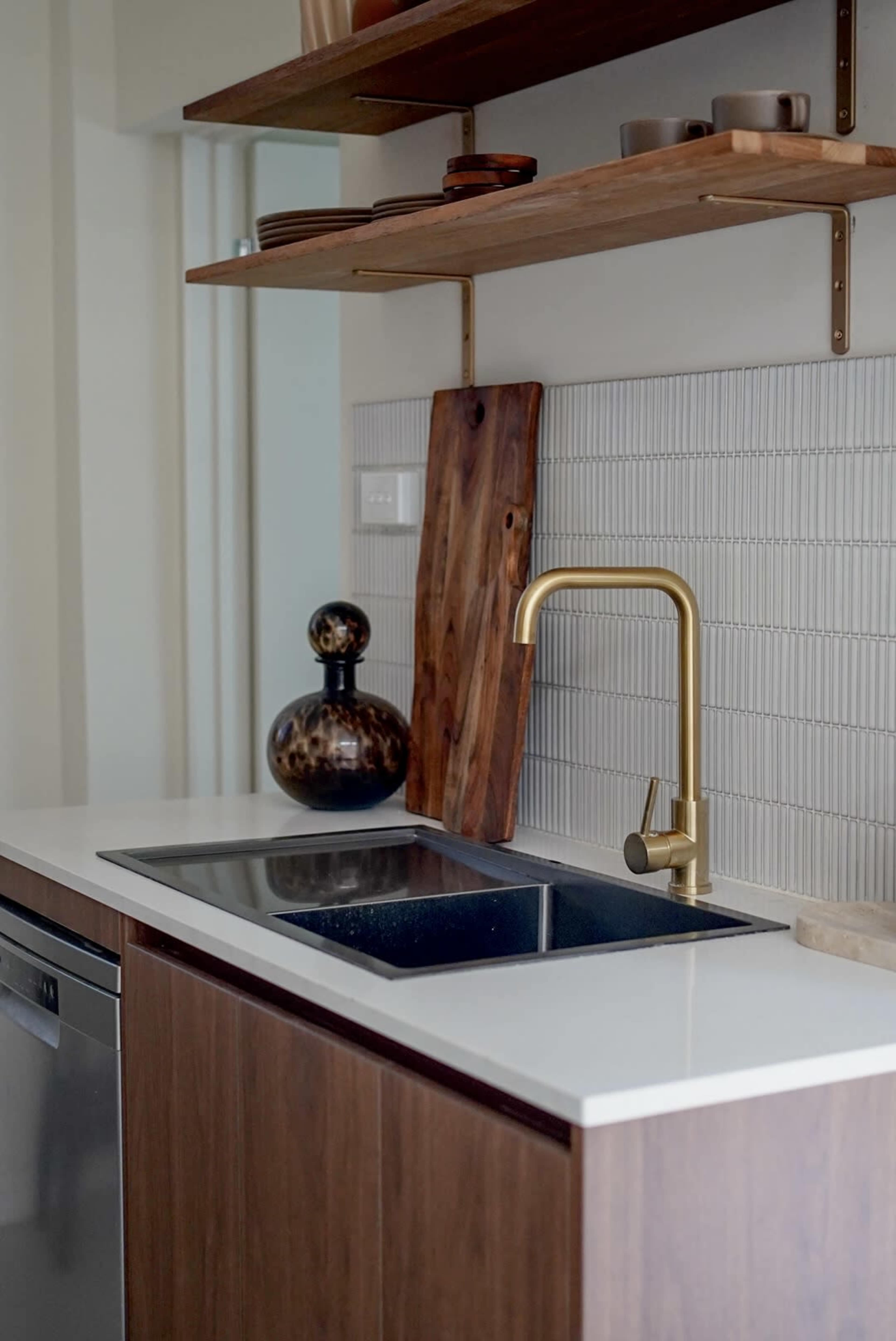A modern kitchen featuring a sleek sink with a gold faucet, wooden shelves, and a textured backsplash.