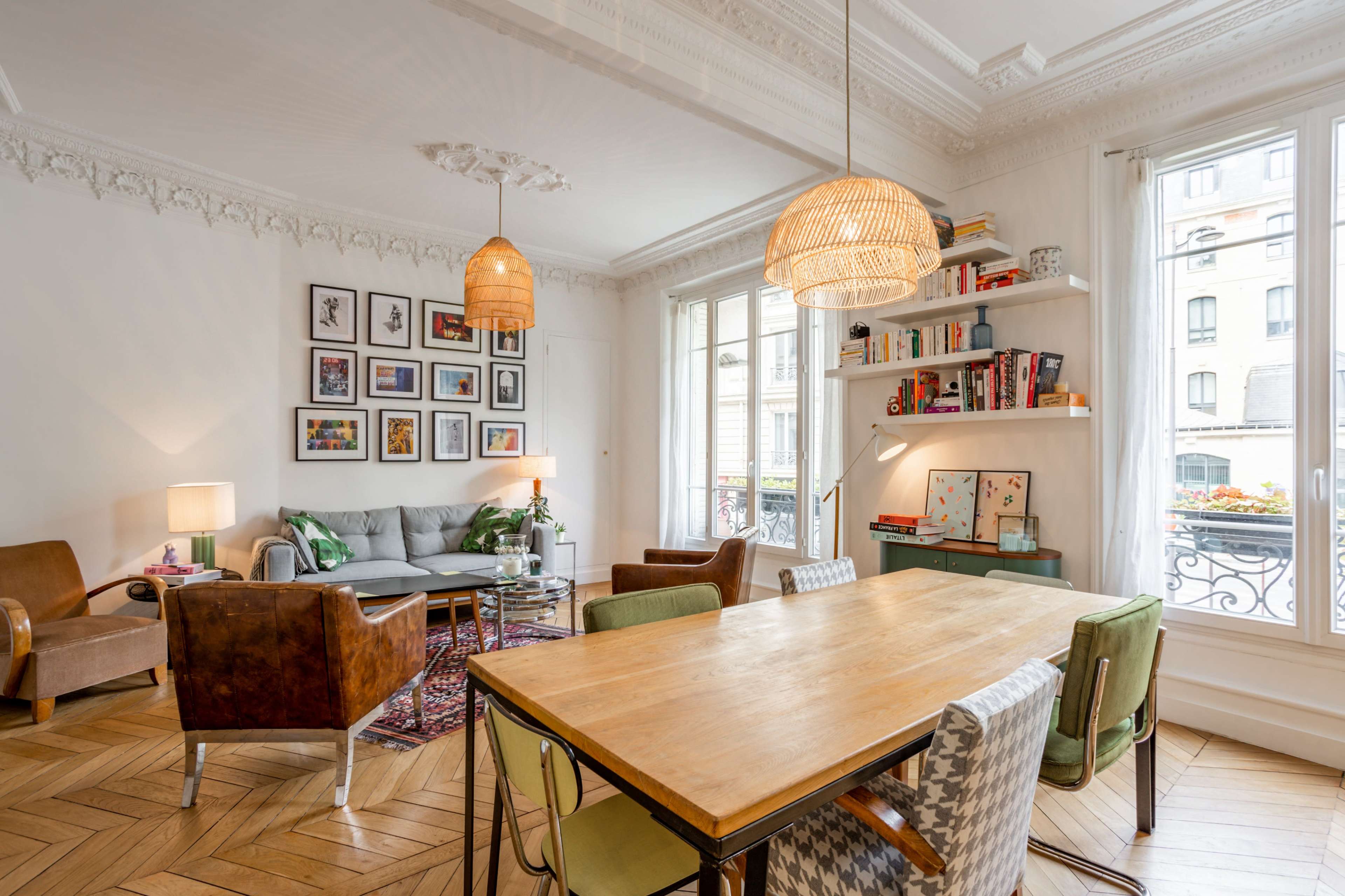 A stylish living area featuring a dining table, comfortable seating, bookshelves, and large windows with decorative molding.