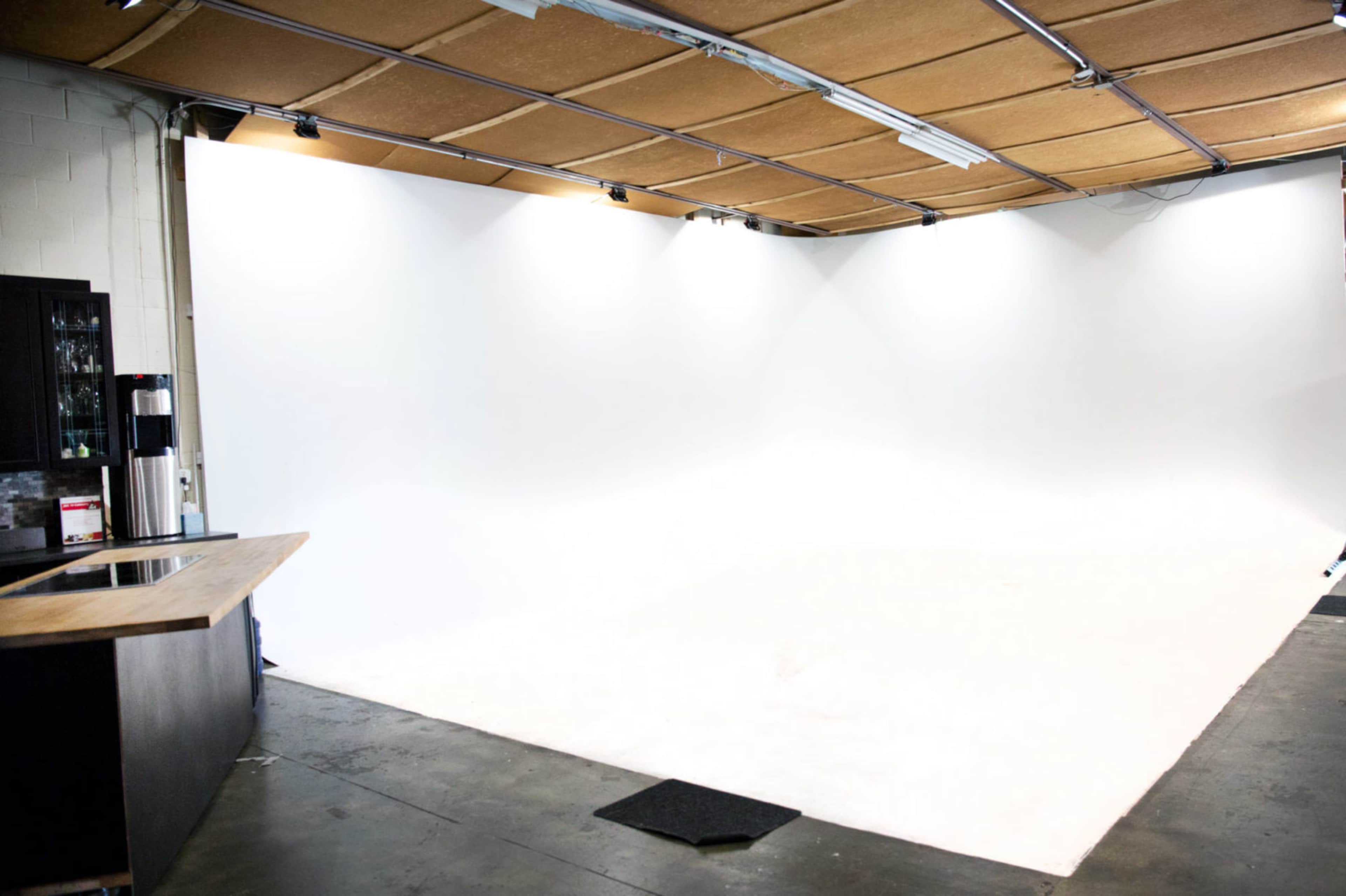 An empty studio space with a white backdrop, a wooden bar counter on the left, and track lighting on the ceiling.