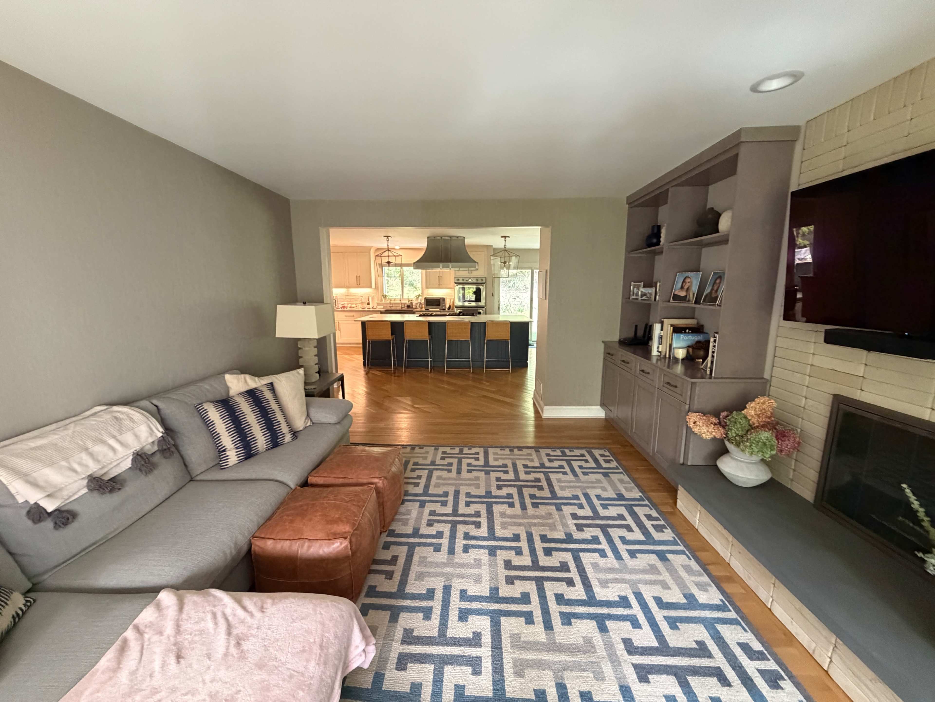 The image shows a modern living room with a gray sectional sofa, a brown leather ottoman, and a patterned rug, leading to an adjacent open kitchen area.