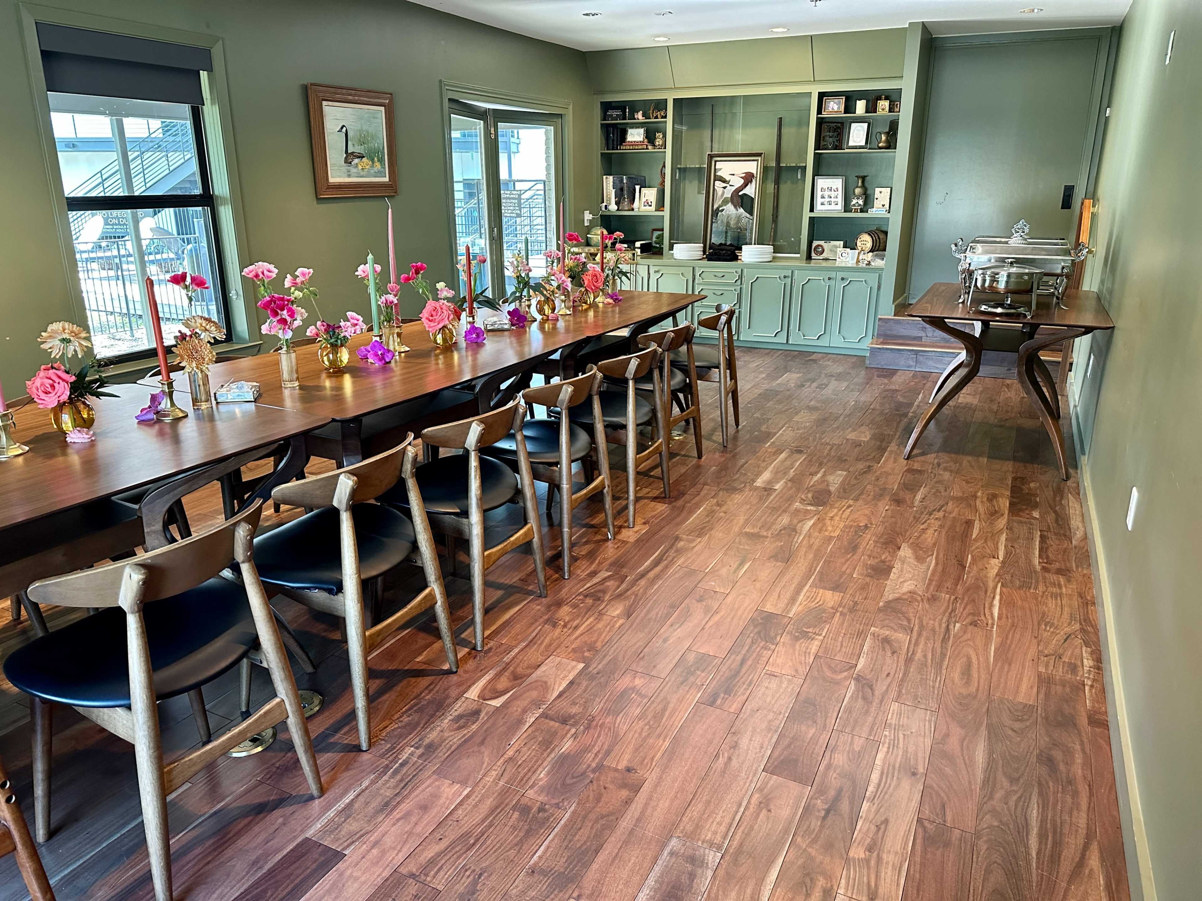 The image shows a spacious dining area with a long wooden table set with floral centerpieces, surrounded by chairs, and a green sideboard against the wall.