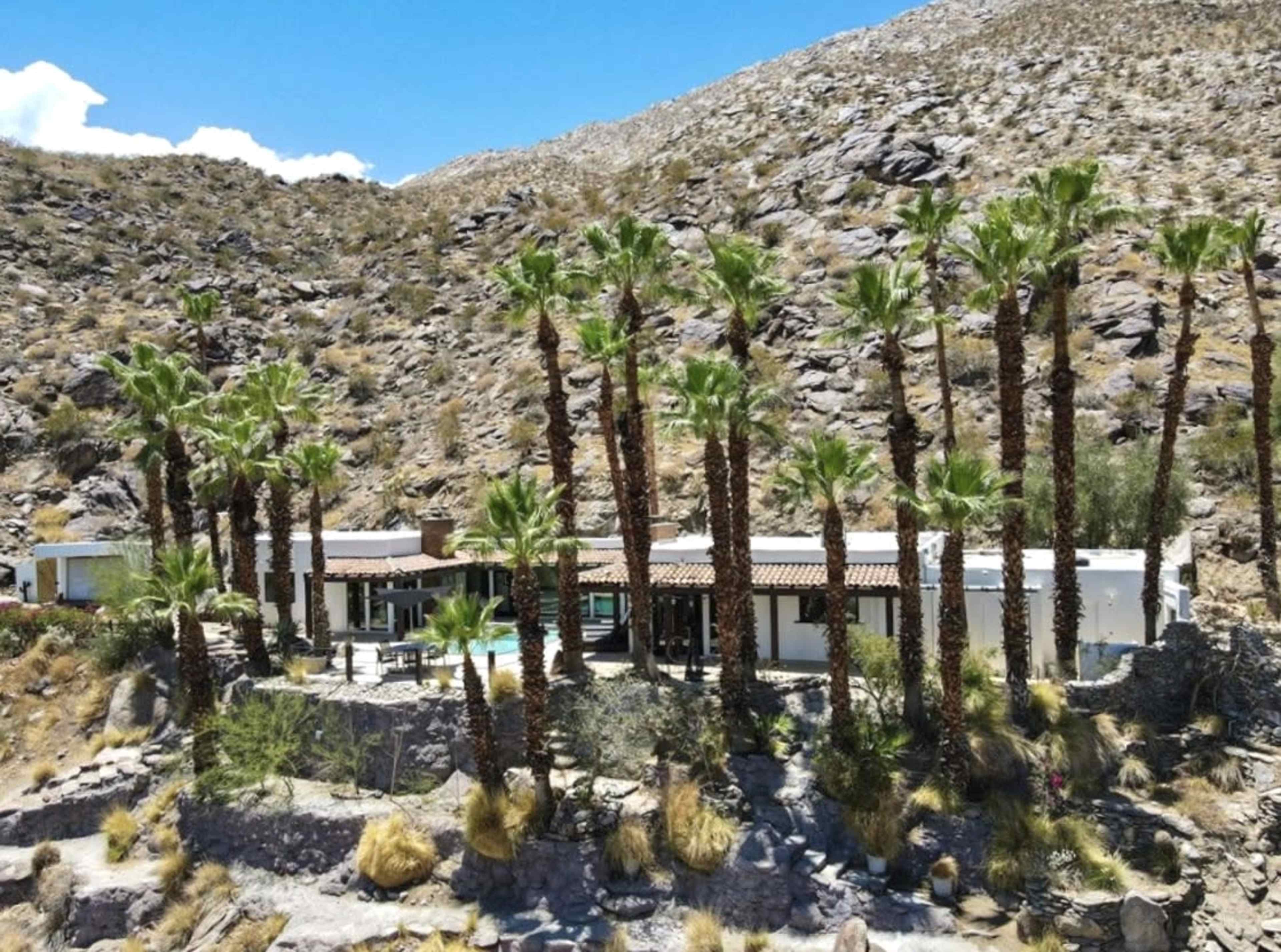 A modern house surrounded by tall palm trees, set against a mountainous, rocky landscape.