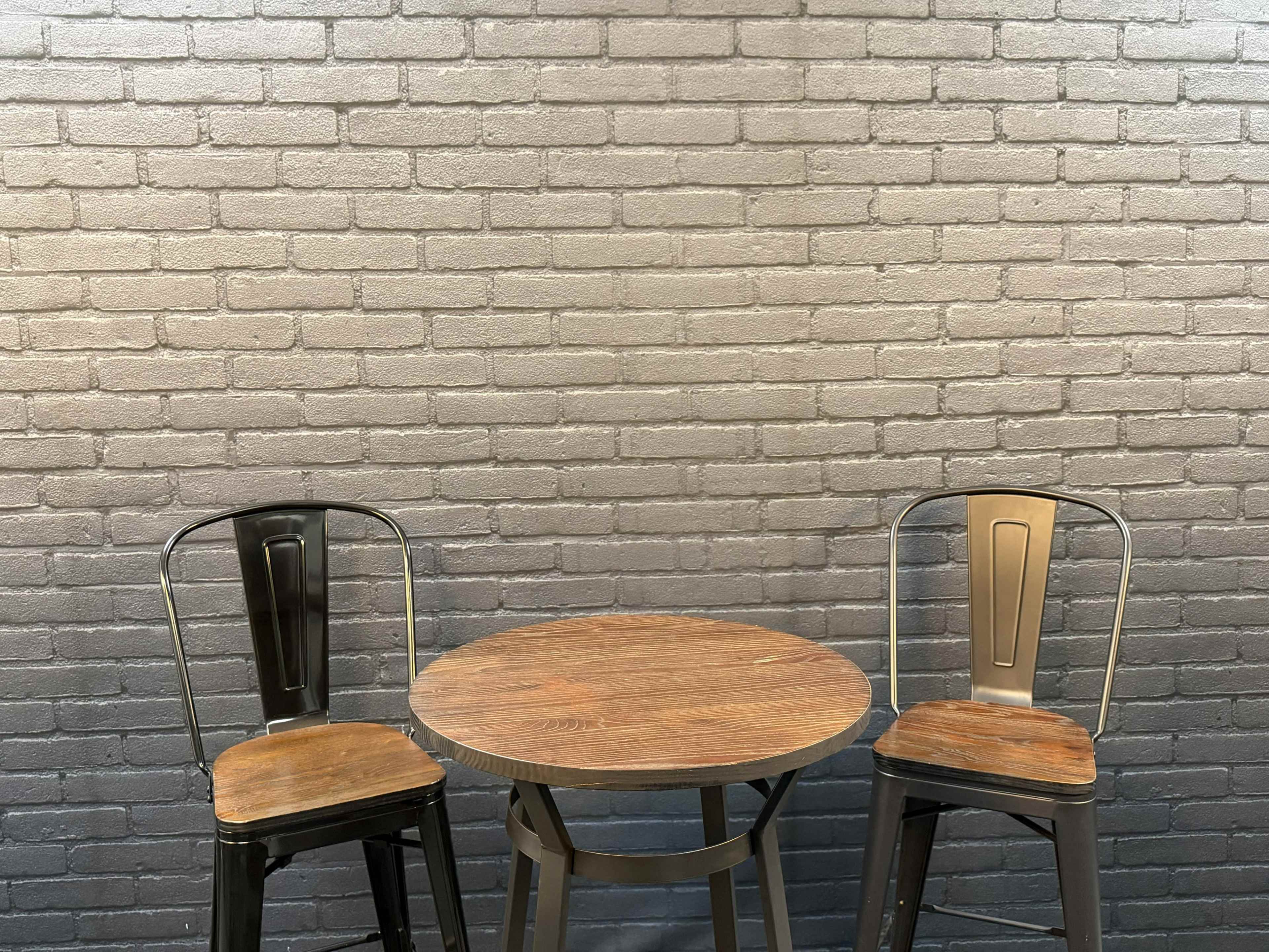 A round wooden table is positioned between two metal stools against a gray brick wall.