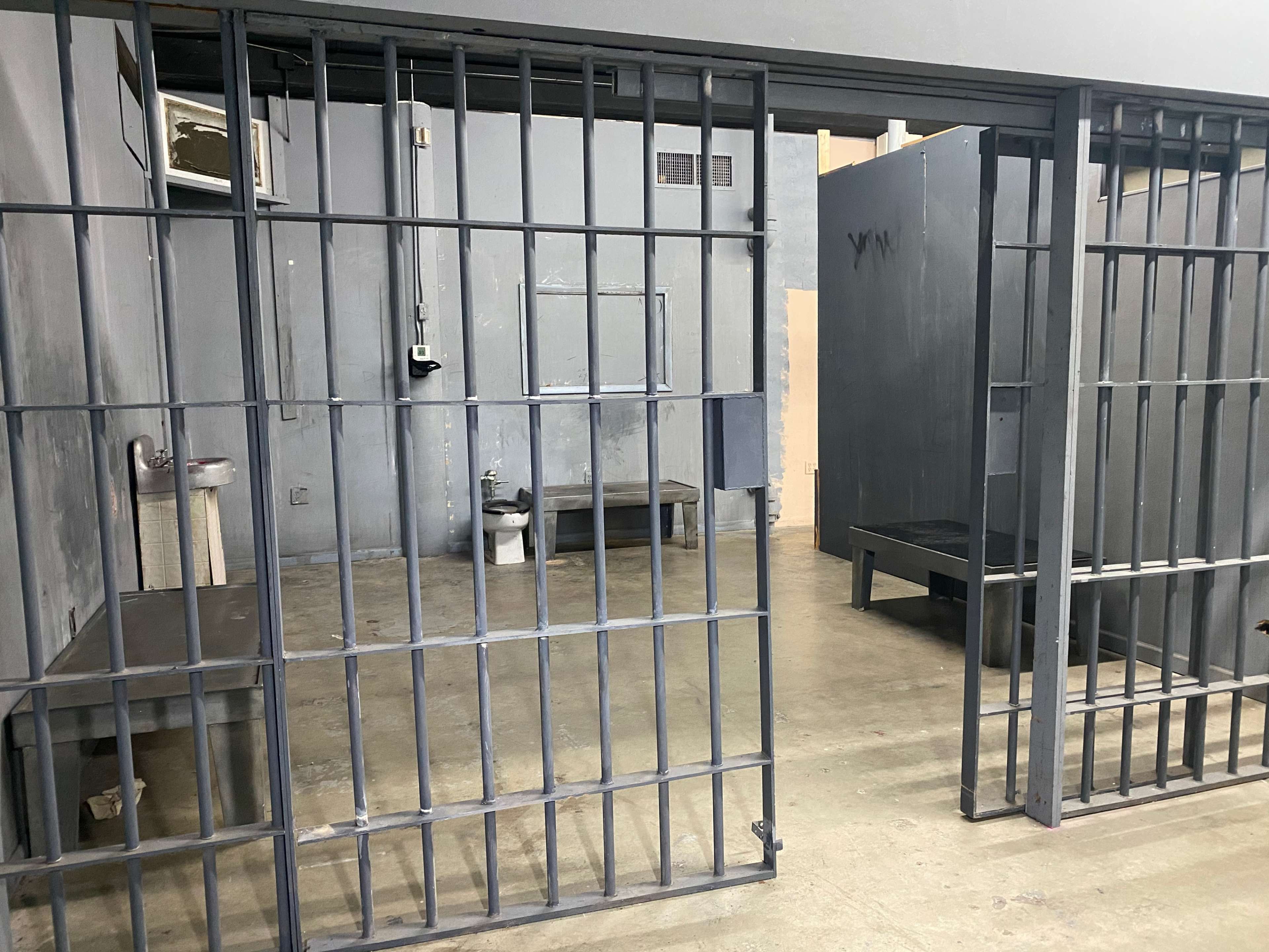 The image shows a partially enclosed area of a prison cellblock with two barred cells and sparse furnishings inside.