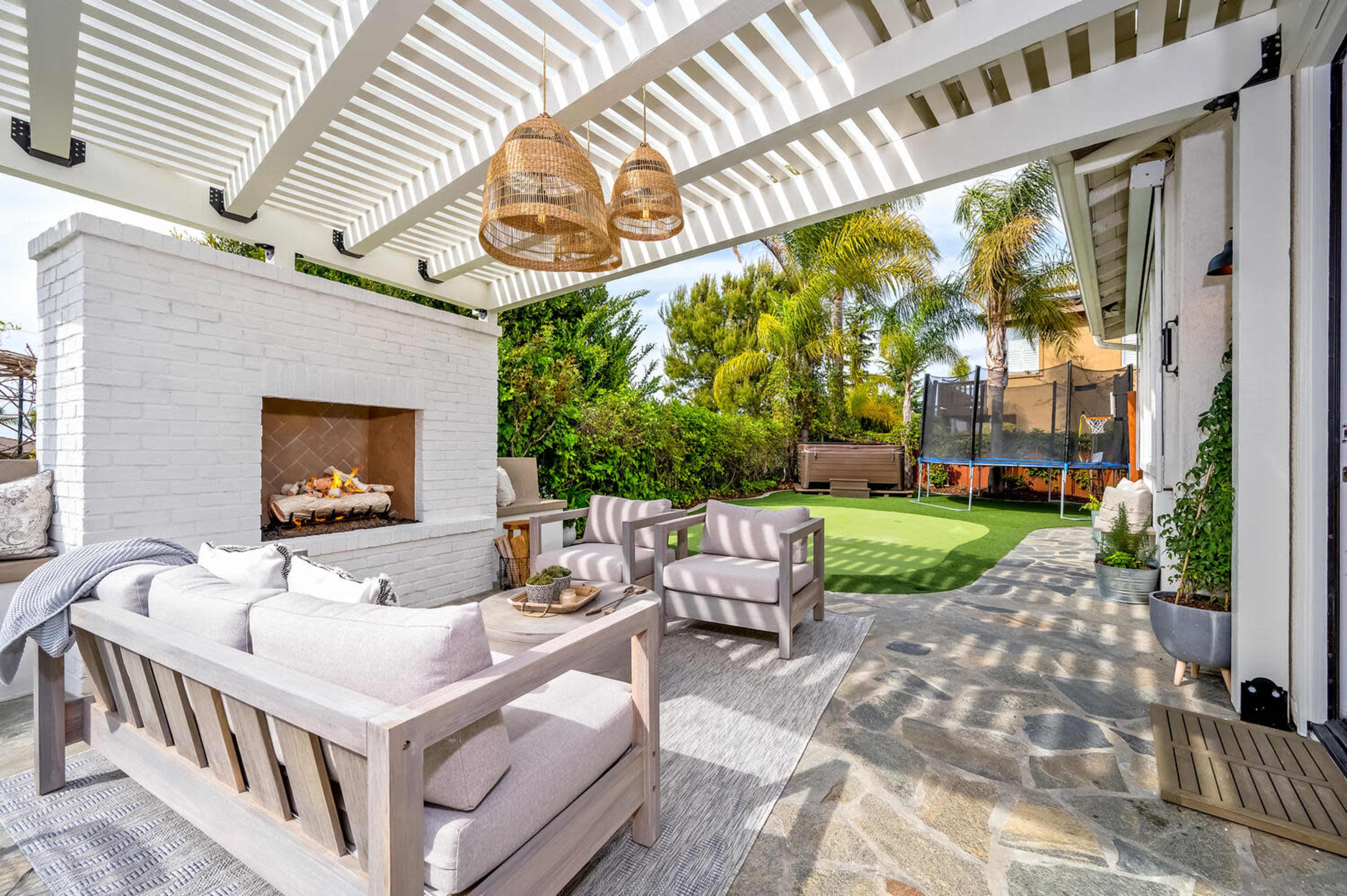 A covered patio features a seating area with wooden furniture, a stone fireplace, and a view of a landscaped garden with palm trees and a trampoline.