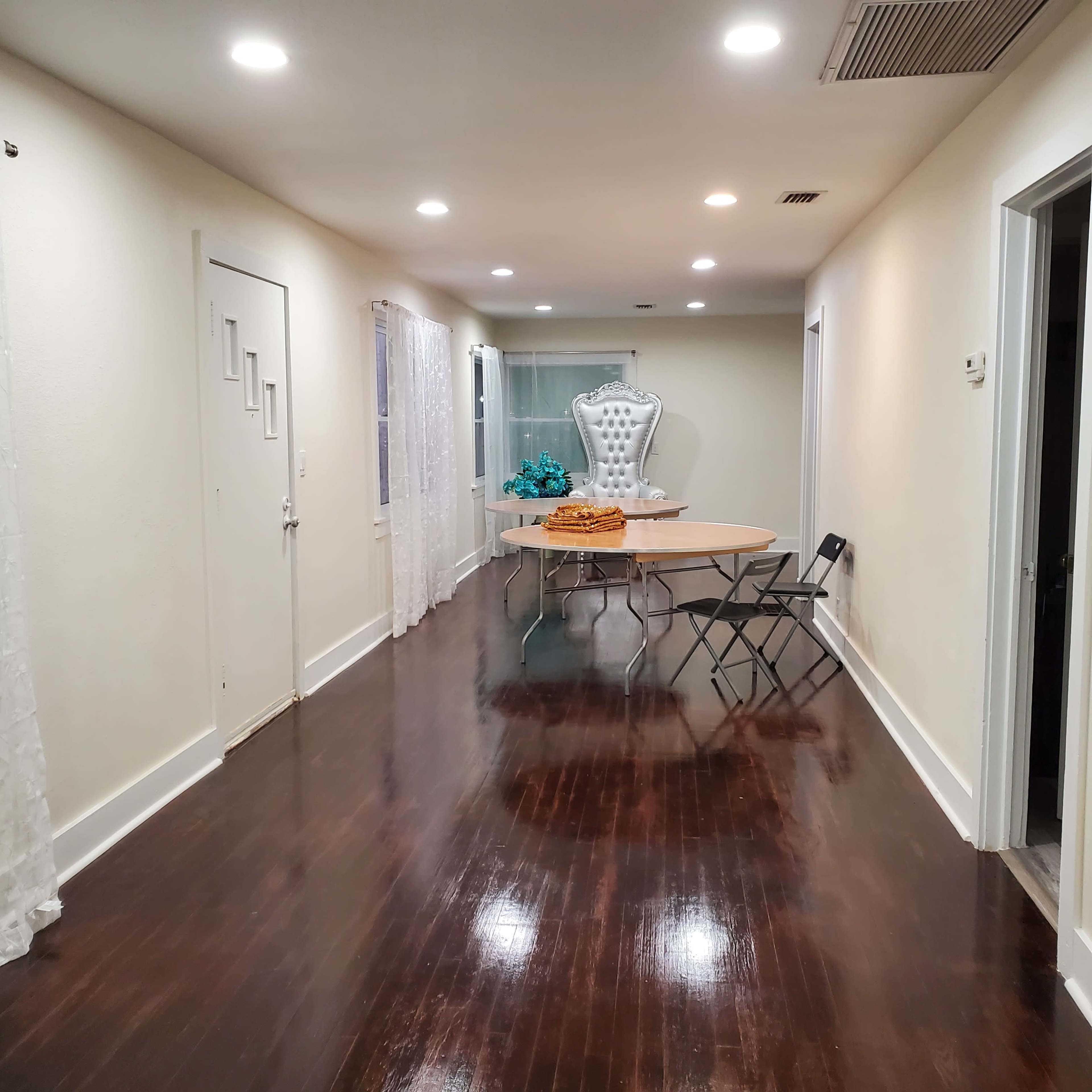 A well-lit hallway features a table with a cake and a chair, leading to a room with a decorative mannequin and sheer curtains.