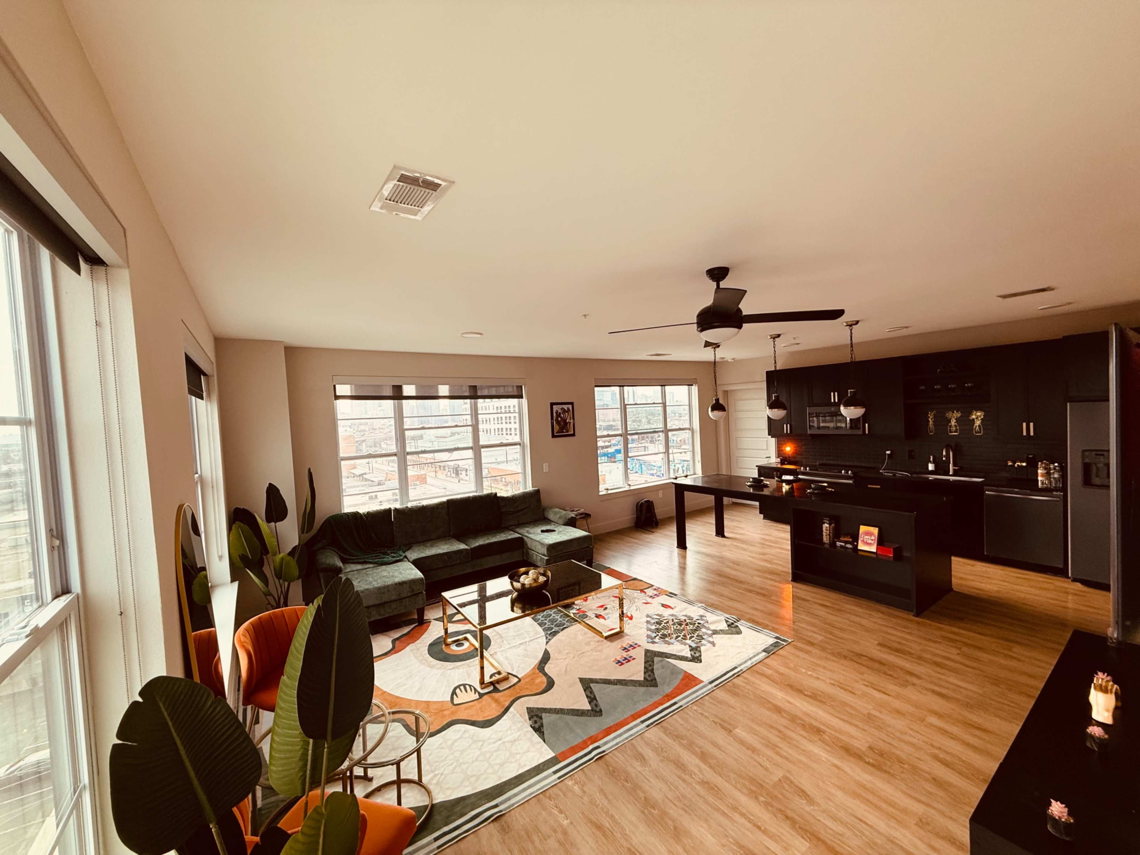The image shows a modern living room with a green sofa, a coffee table, and large windows overlooking an urban view, along with a stylish kitchen area in the background.