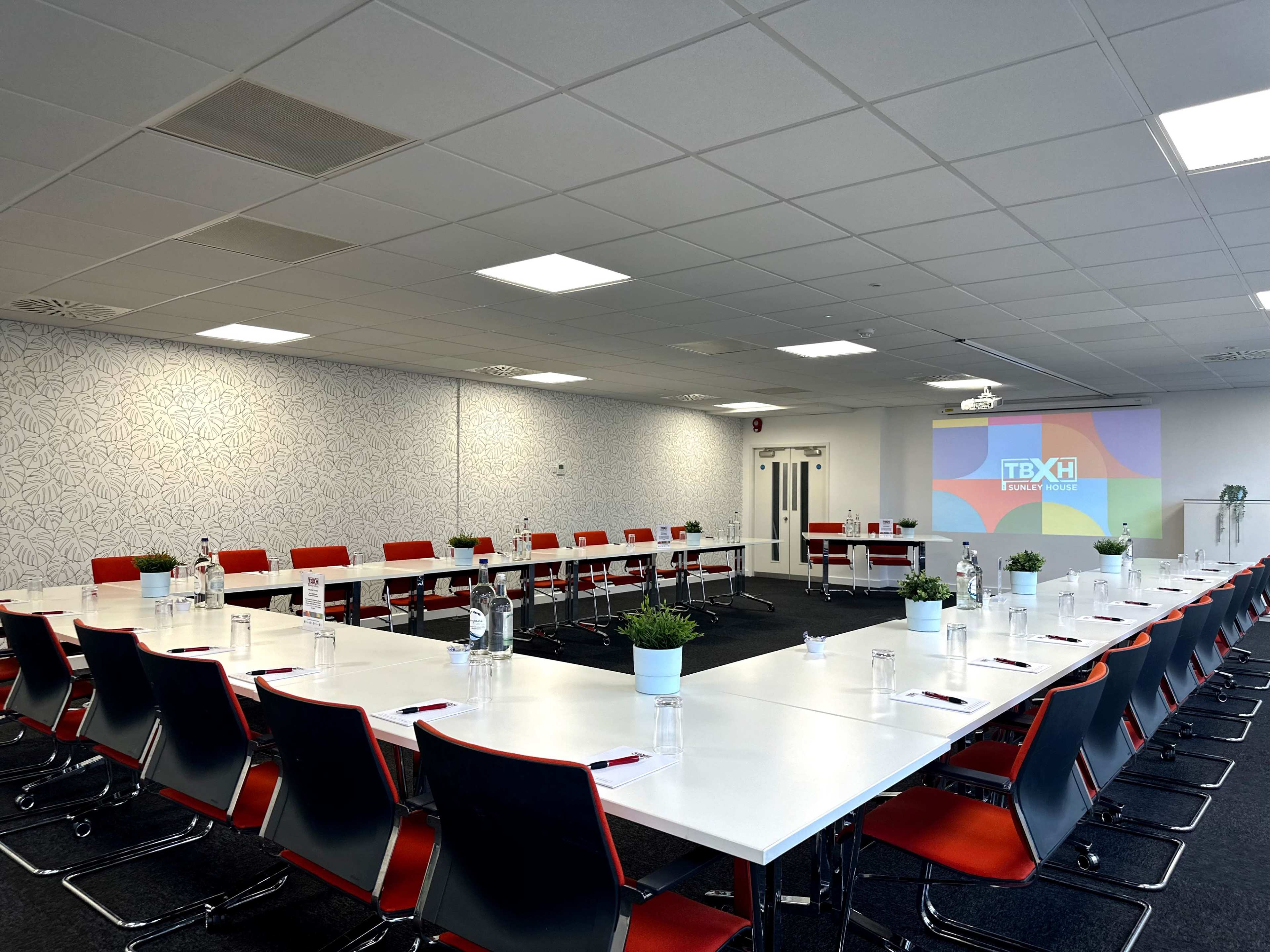A spacious conference room features a large, U-shaped arrangement of white tables and red chairs, with a projector screen displaying a logo.