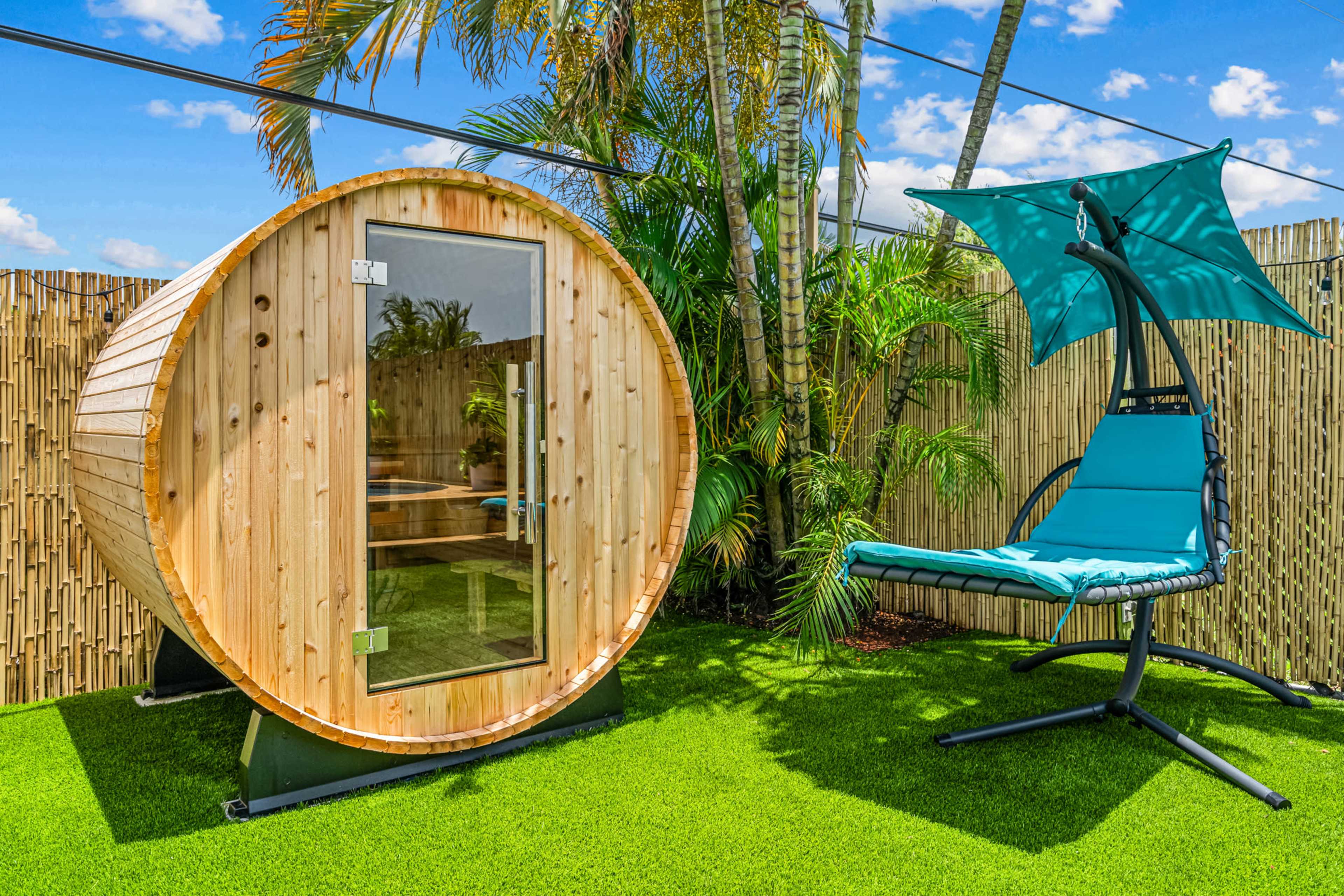A wooden sauna in a circular shape sits beside a teal hanging chair on a green artificial lawn, surrounded by tropical plants and a bamboo fence.
