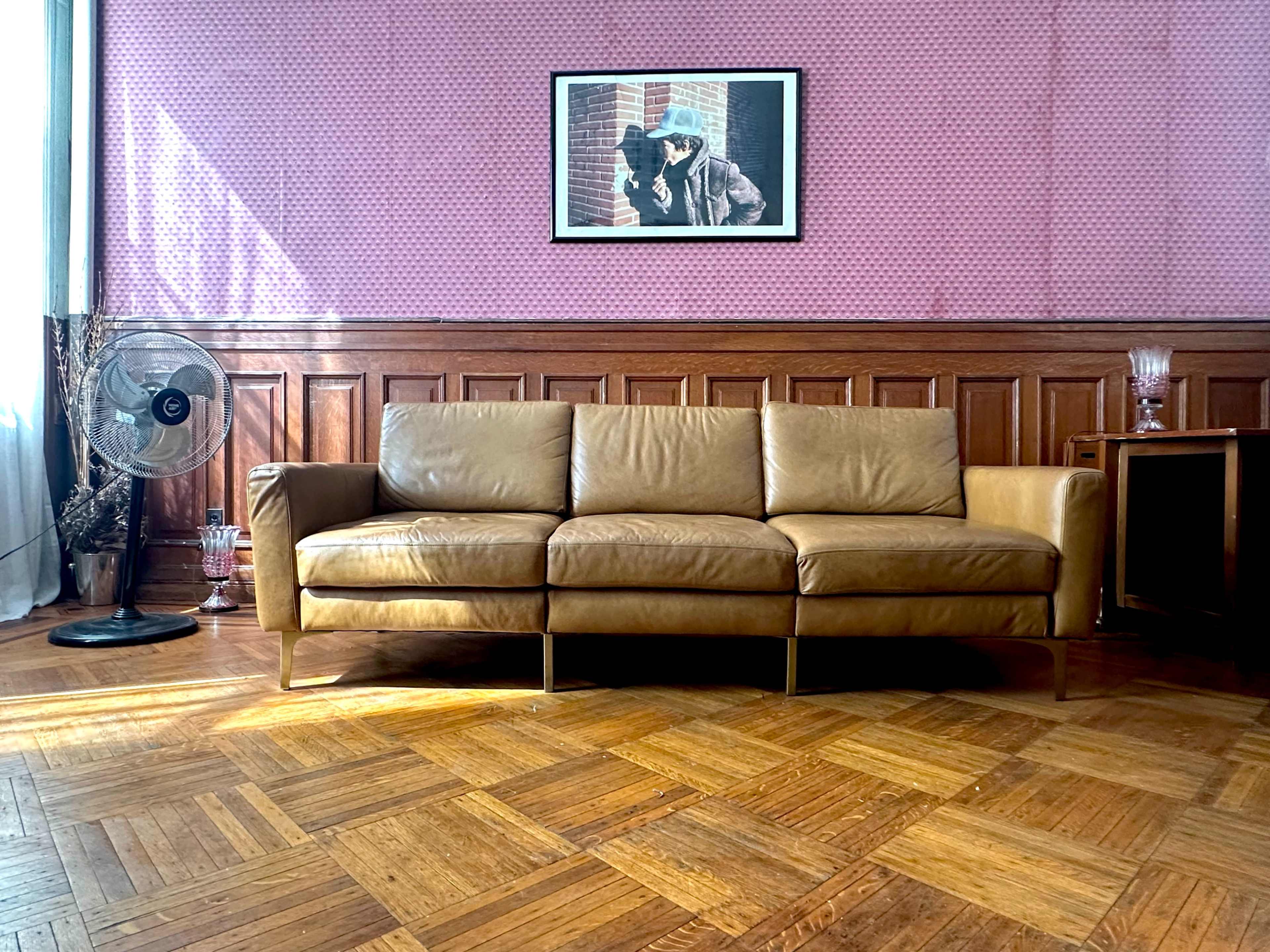 The image shows a brown leather sofa in a room with patterned pink wallpaper, a wooden floor, a vintage fan, and a framed photo on the wall.