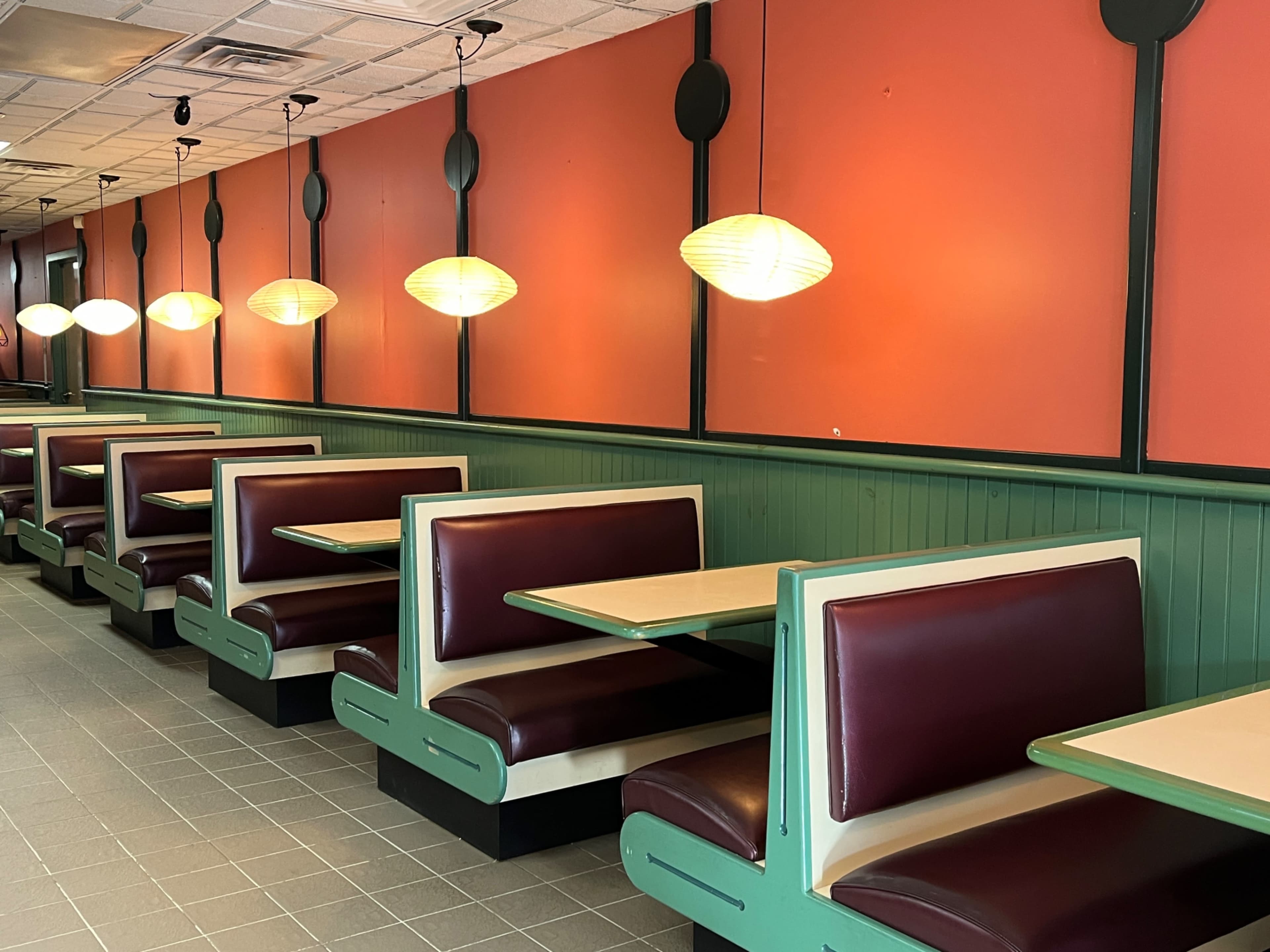 A diner interior with red walls, green paneling, and several booths arranged along the space, illuminated by hanging pendant lights.