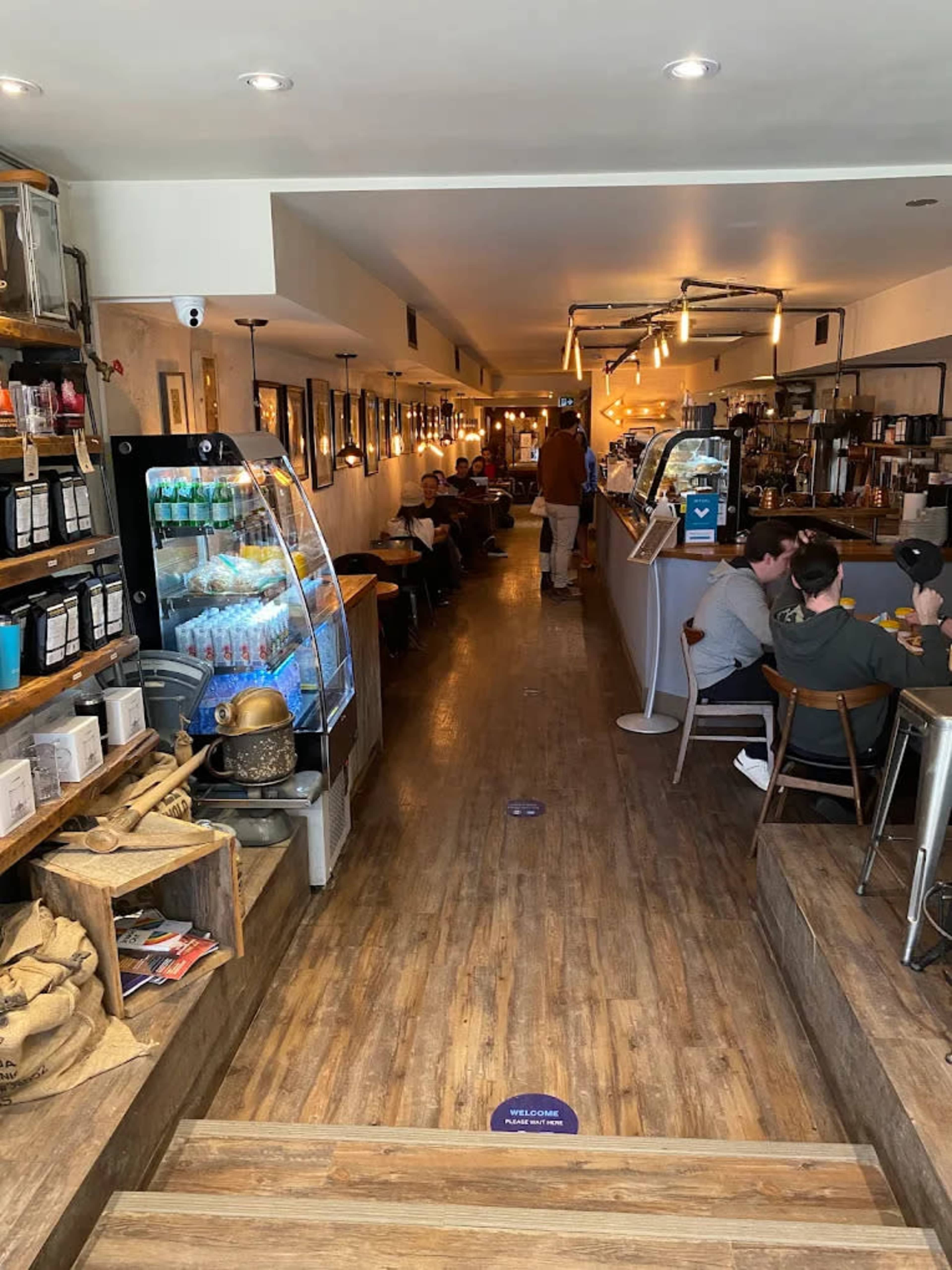 The image shows the interior of a café with a long wooden corridor, seating on both sides, and a counter at the end where drinks are served.