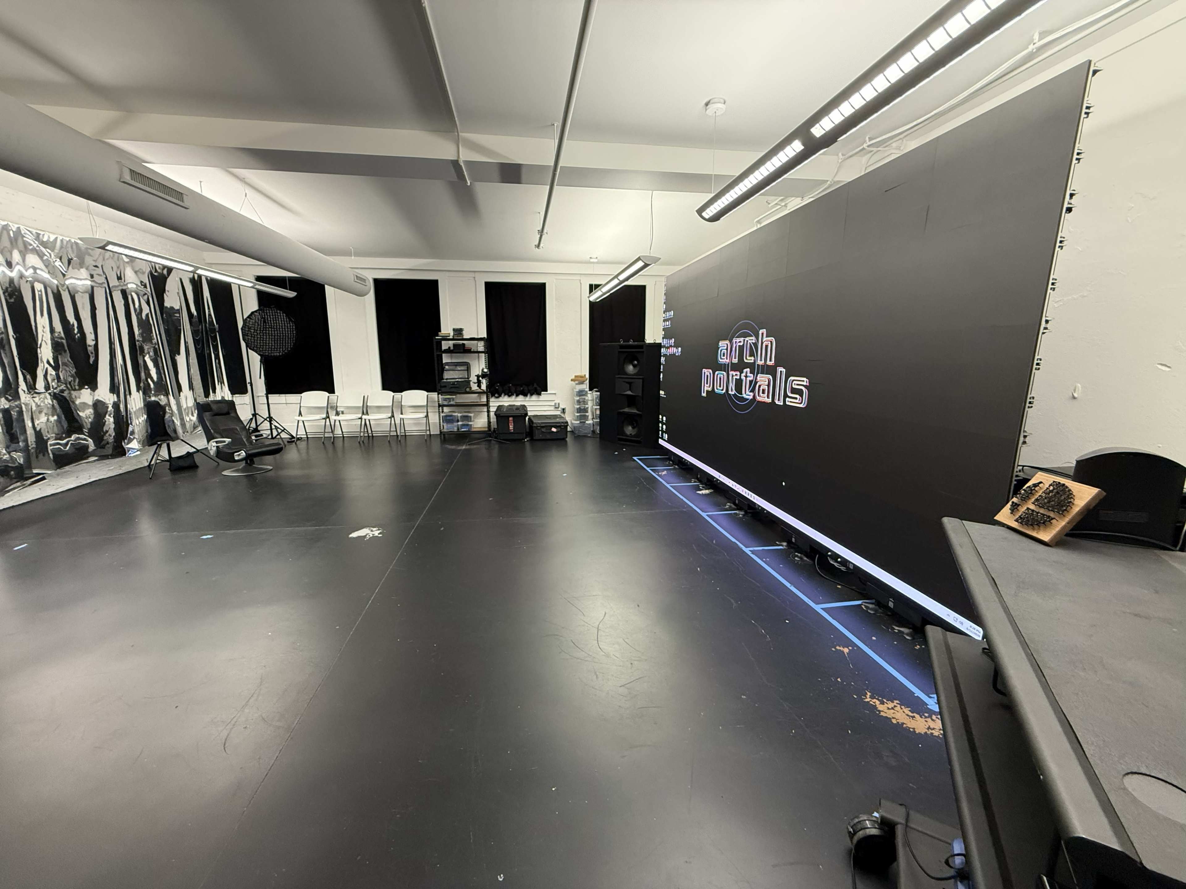 The image shows a spacious studio with a large dark backdrop featuring the text "arch portals," empty chairs lined up on one side, and various equipment visible in the background.