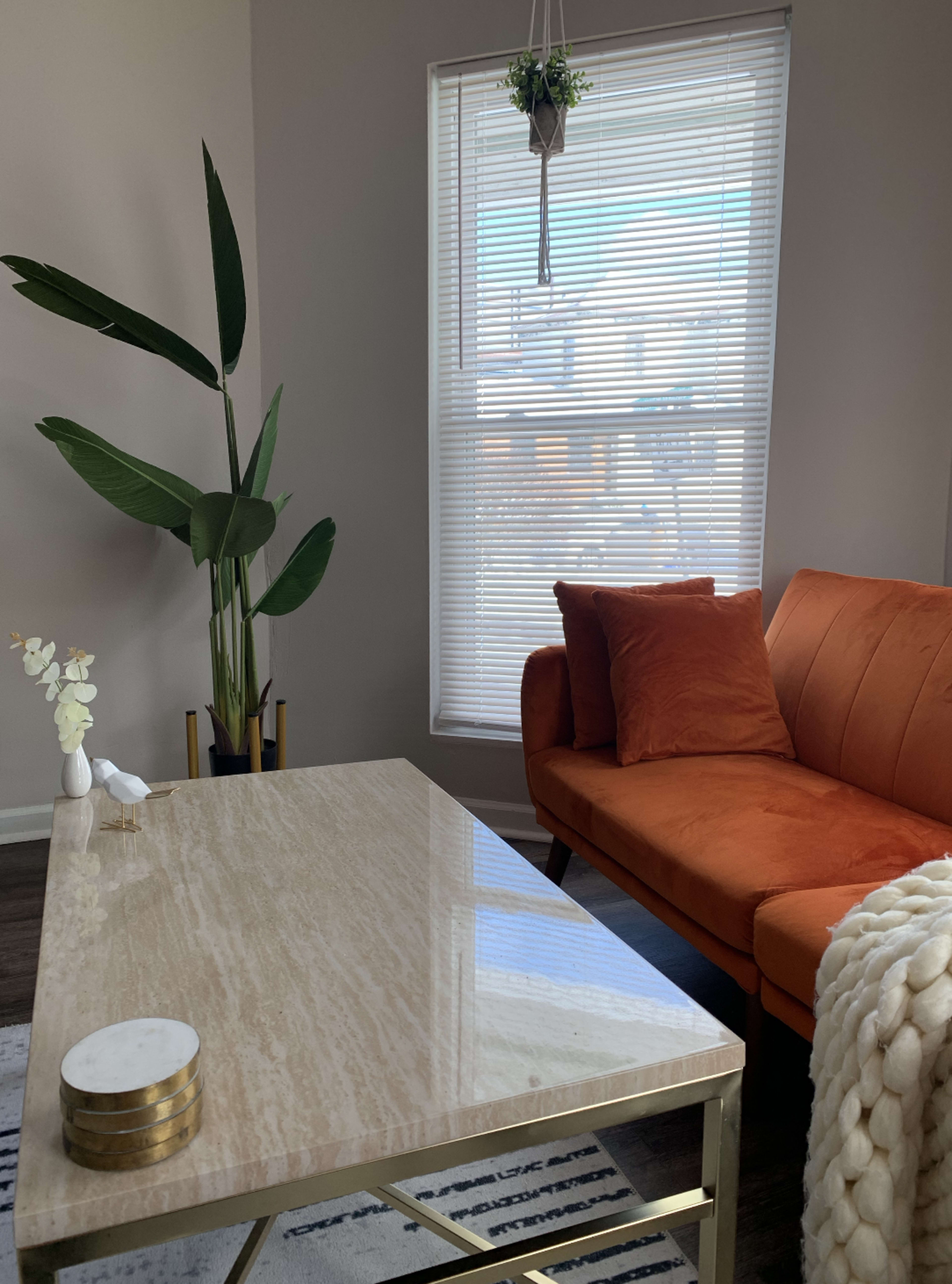 A living room with an orange sofa, a marble coffee table, a potted plant, and a window with blinds.