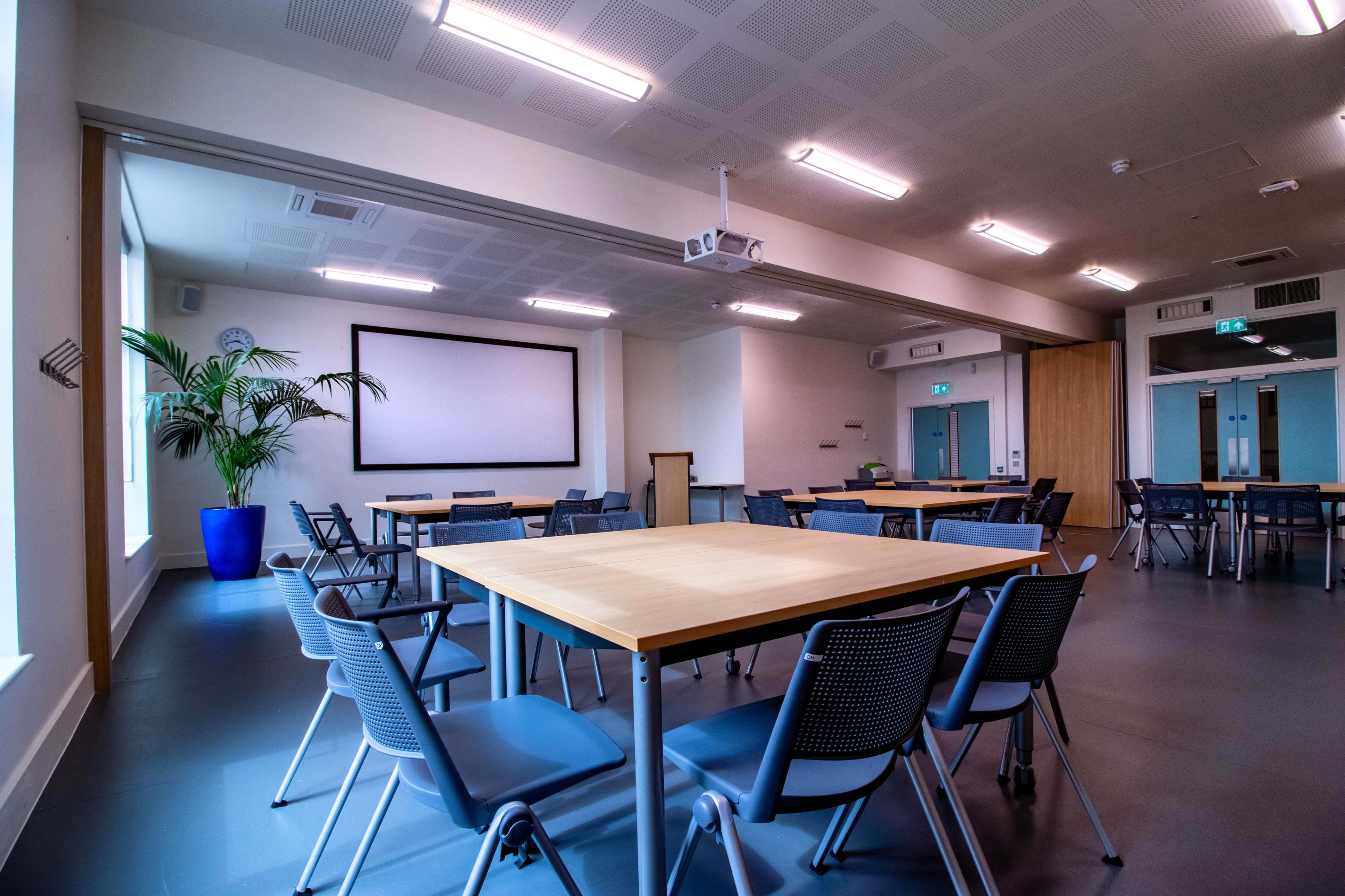 A spacious, well-lit meeting room with several wooden tables and chairs arranged around a central area, featuring a large projection screen on one wall.